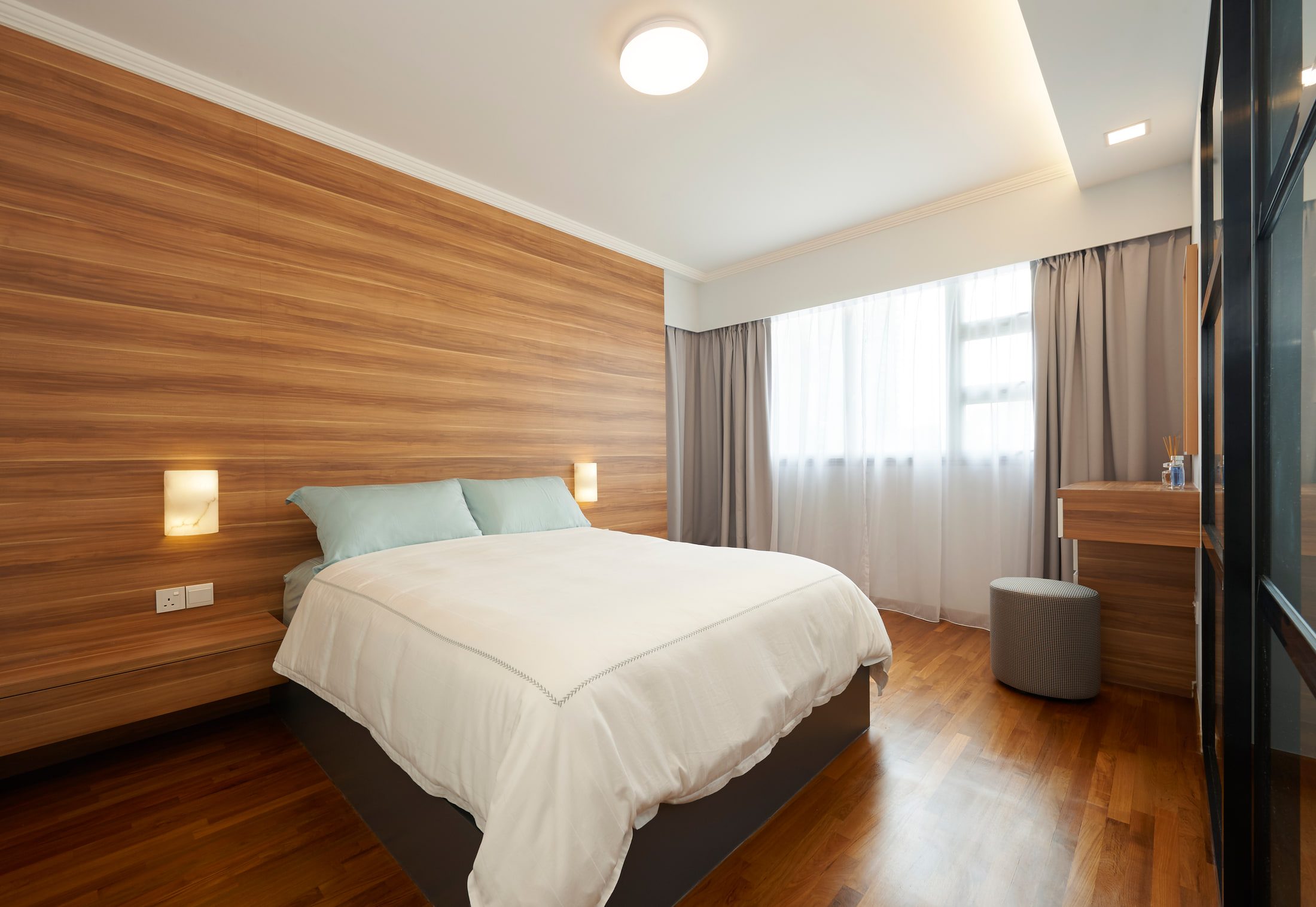 The same bedroom seen from the doorway, alabaster wall lights recessed into the timber panelling and grey curtains drawn back from a full-width window.