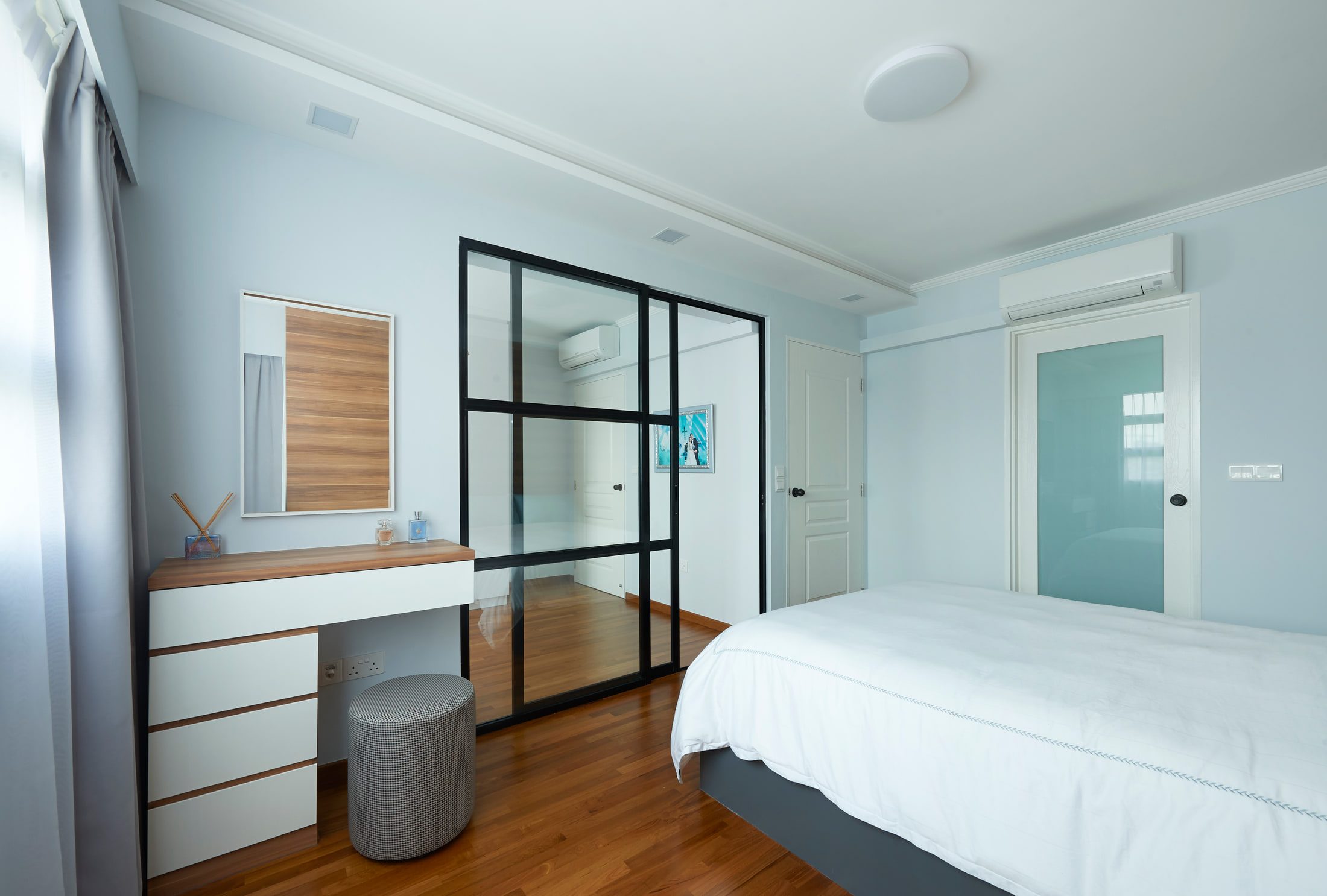 A bedroom in pale blue with a timber-topped dressing table, a chest of drawers and a black-framed glazed partition beside the bed.