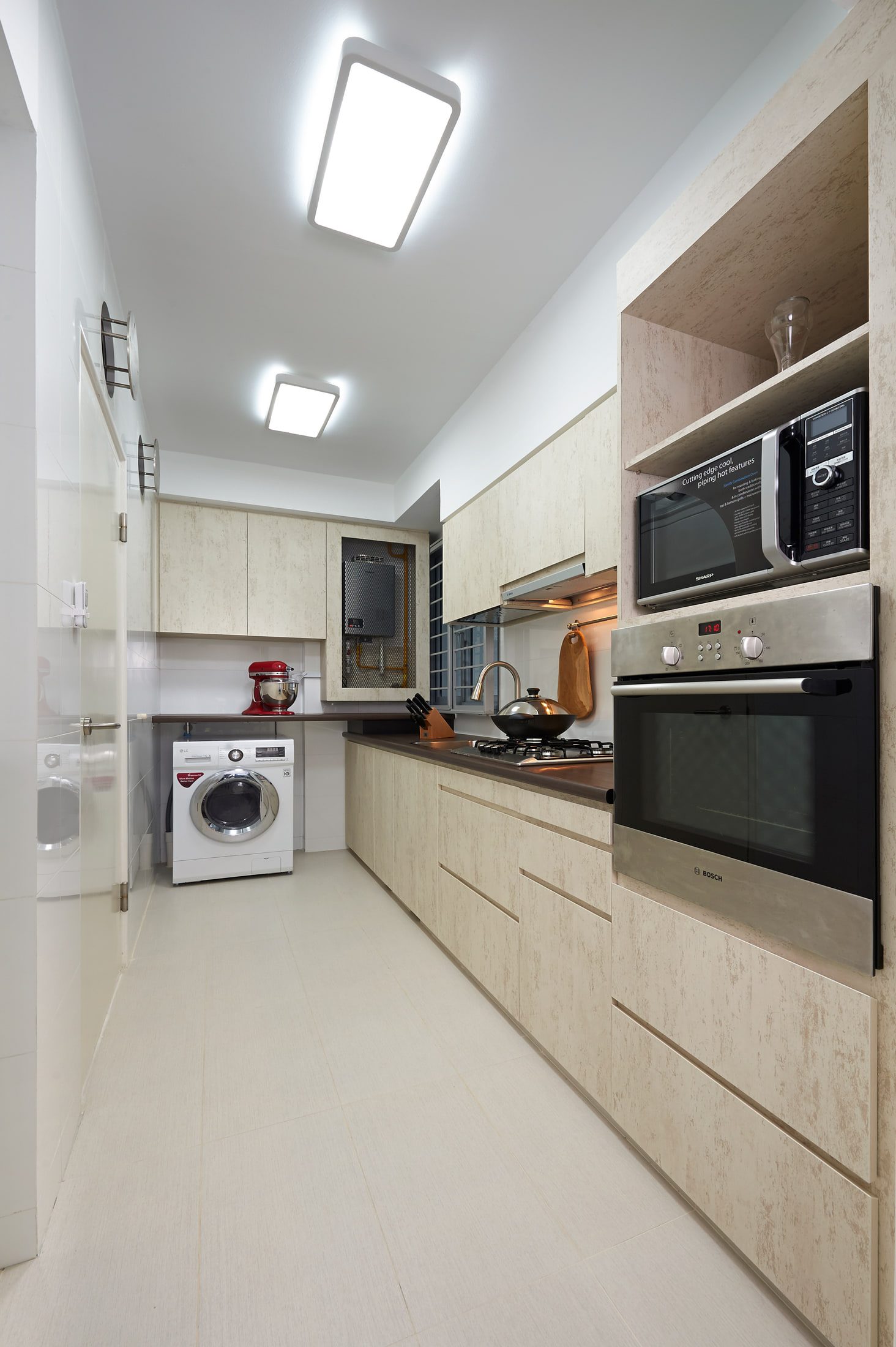 A narrow galley kitchen with travertine-patterned laminate fronts, a dark worktop, a wall oven and microwave stack, and a washing machine set into the run beyond a glazed door.