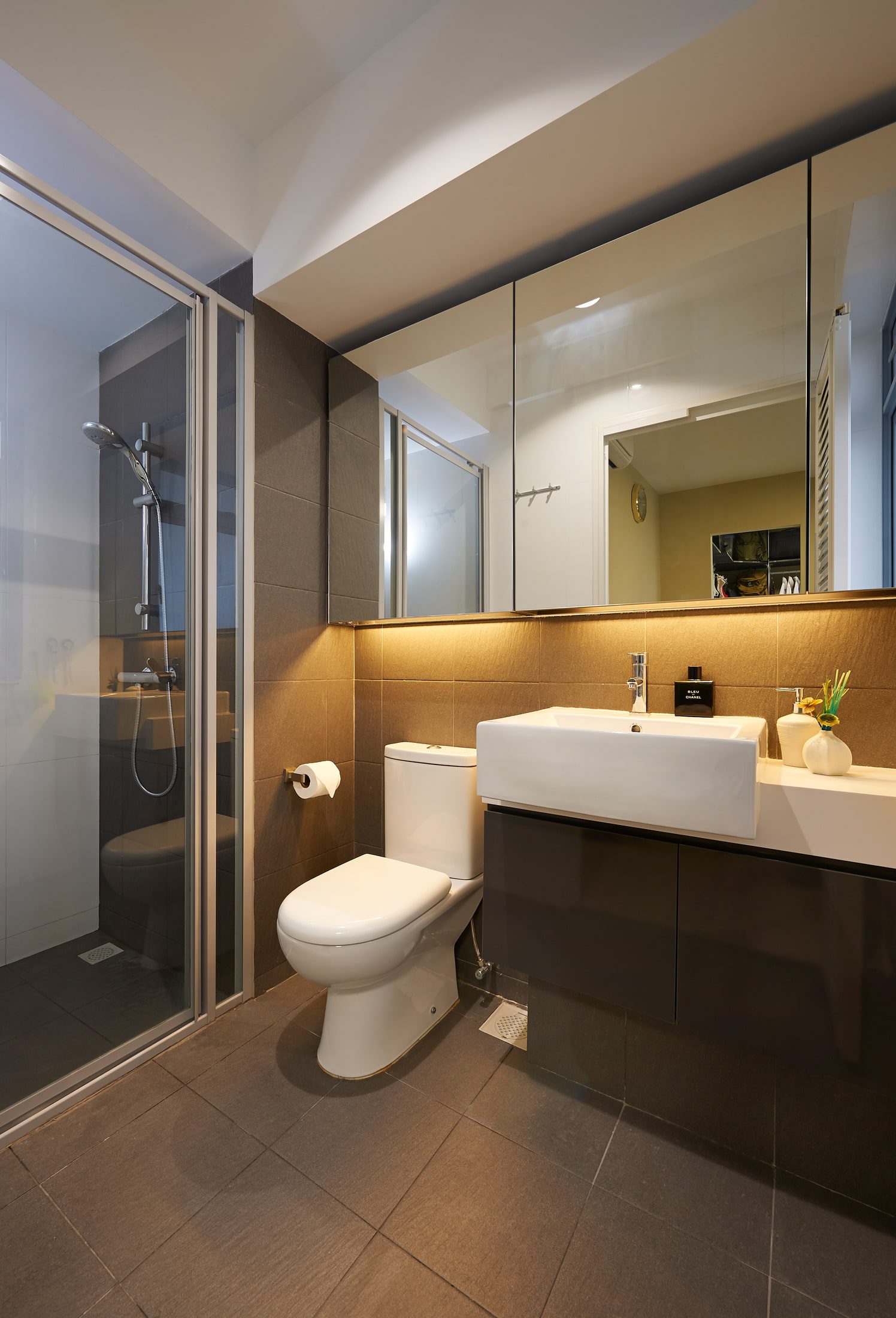 A bathroom with a rectangular vessel basin on a dark gloss vanity, a bronze-toned tile band lit from under the mirror cabinets, and a framed shower enclosure in charcoal tile.
