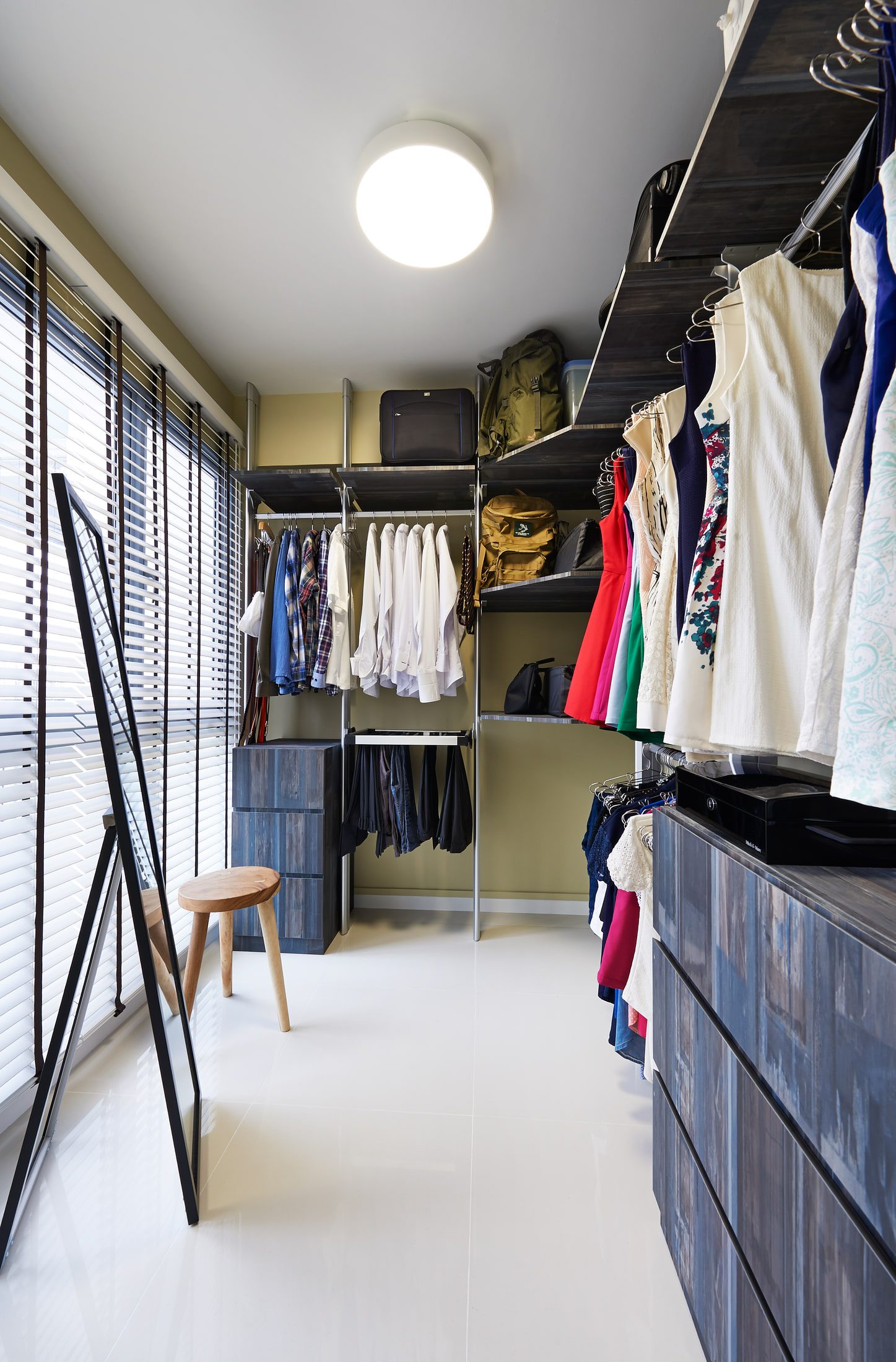 A walk-in wardrobe with open steel-post shelving in dark timber, hanging rails of shirts and dresses, a leaning full-length mirror and a round-topped stool by the venetian blinds.