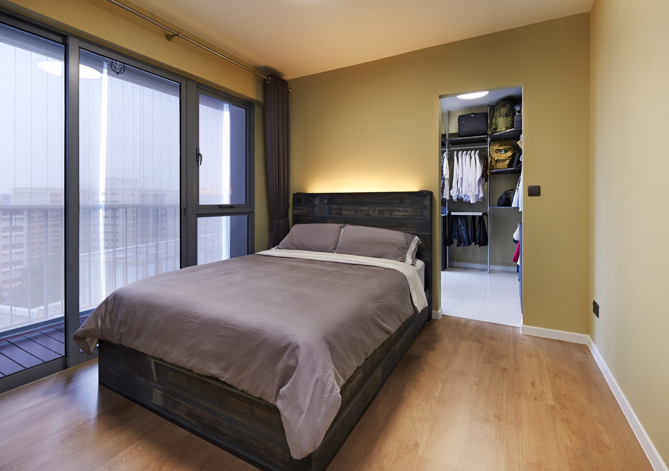 A bedroom in olive with a reclaimed-timber bed frame and headboard lit from above, sliding glazing to a narrow balcony, and an open doorway to a lit walk-in wardrobe.