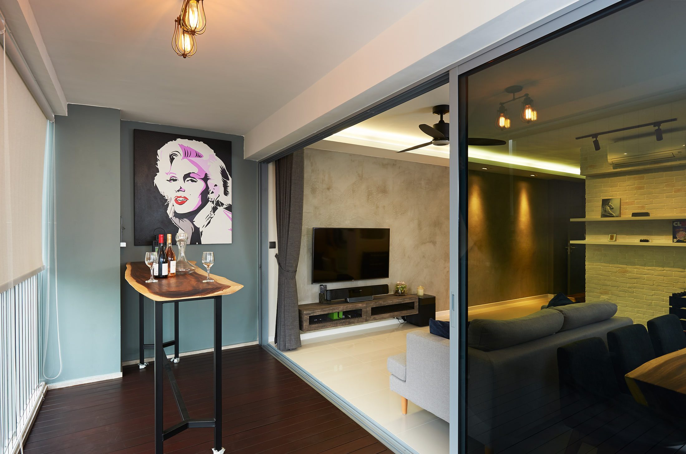 A covered balcony floored in dark timber decking with a live-edge standing bar holding wine and a decanter, a pop-art portrait canvas on a slate-blue wall, and caged pendants overhead.
