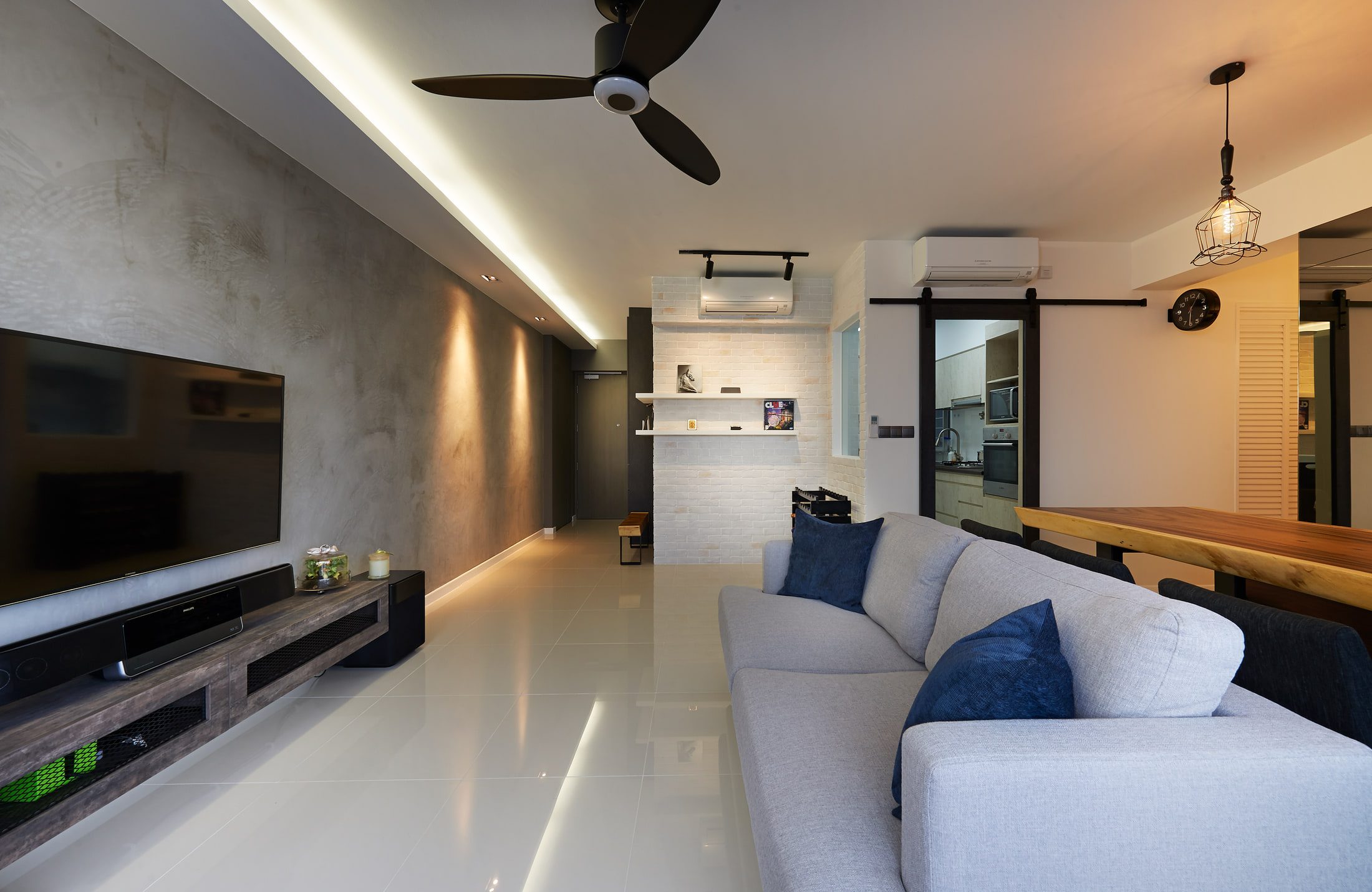 A living room floored in white tile with a grey linen sofa, a cement-rendered wall washed by downlights, a black ceiling fan, and a barn door on an exposed track opening to the kitchen.