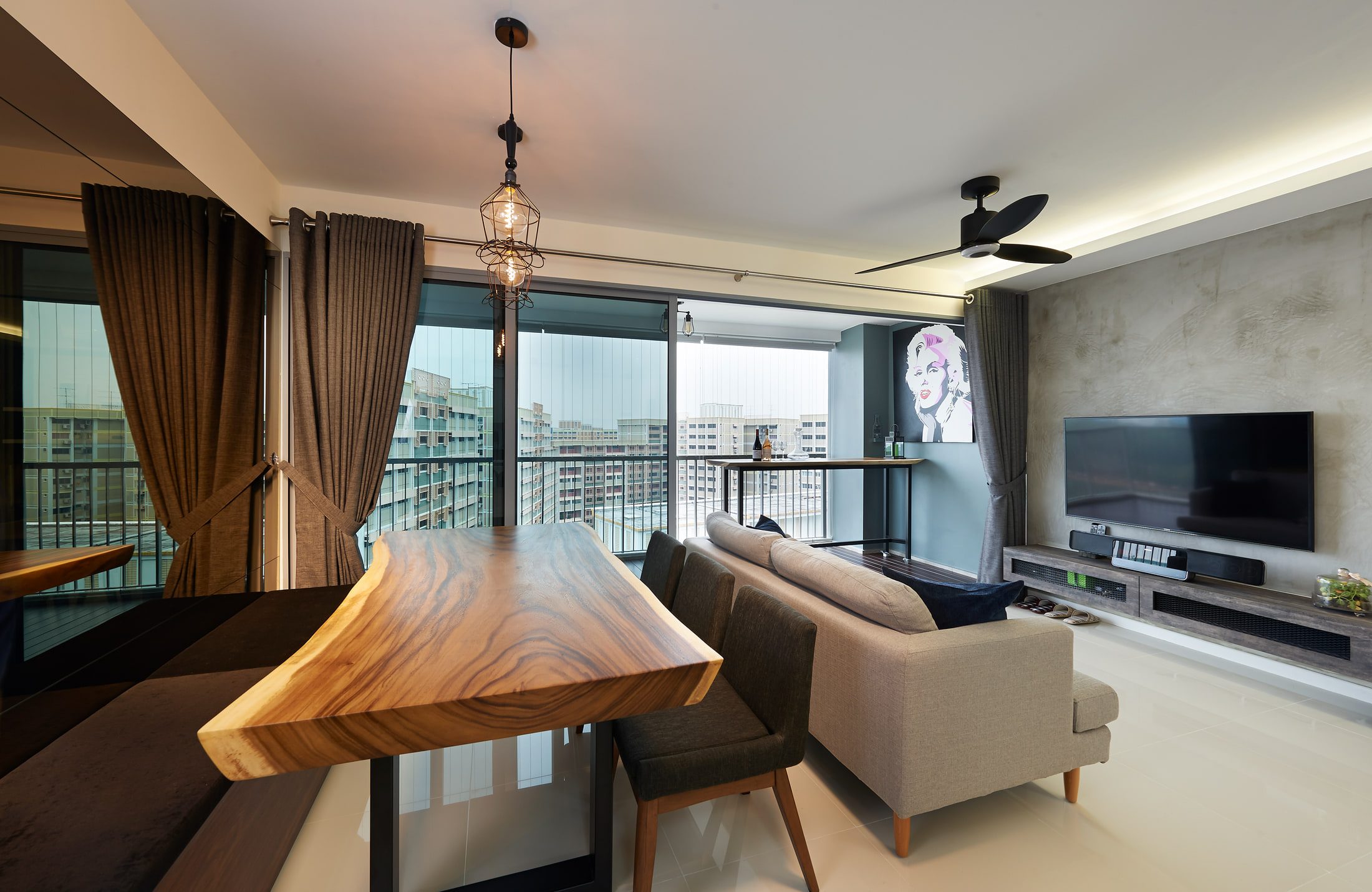 A live-edge hardwood slab table on a black steel frame in front of full-height balcony glazing, with caged filament pendants above and a grey sofa facing a cement-rendered television wall.