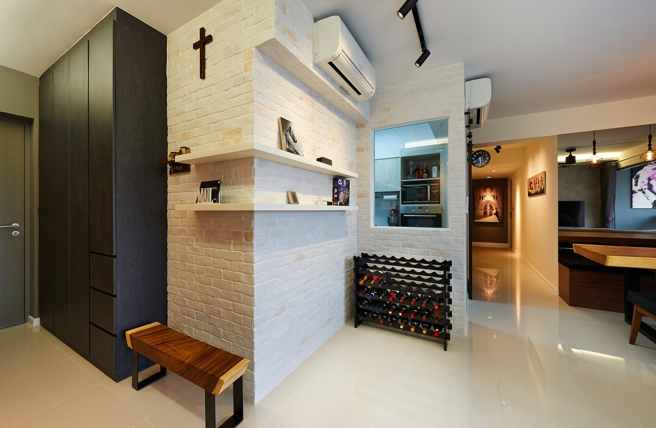 An entrance hall with a whitewashed brick pier carrying two white shelves and a small timber cross, a dark full-height shoe cupboard, a live-edge bench and a black wine rack.