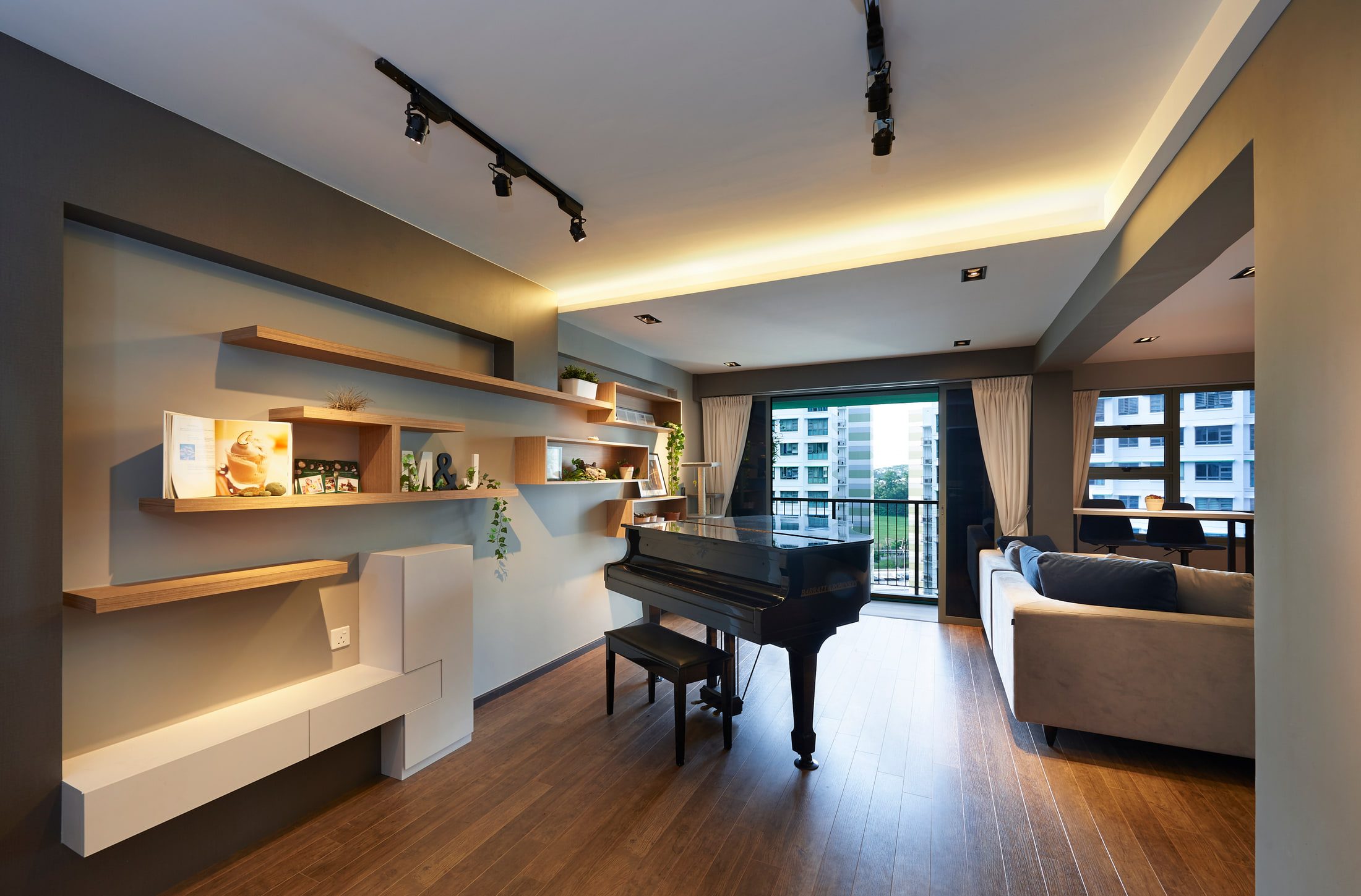 A living room with a black grand piano standing against a balcony window, an asymmetric wall of oak shelves and boxes opposite, and a low white sideboard beneath.