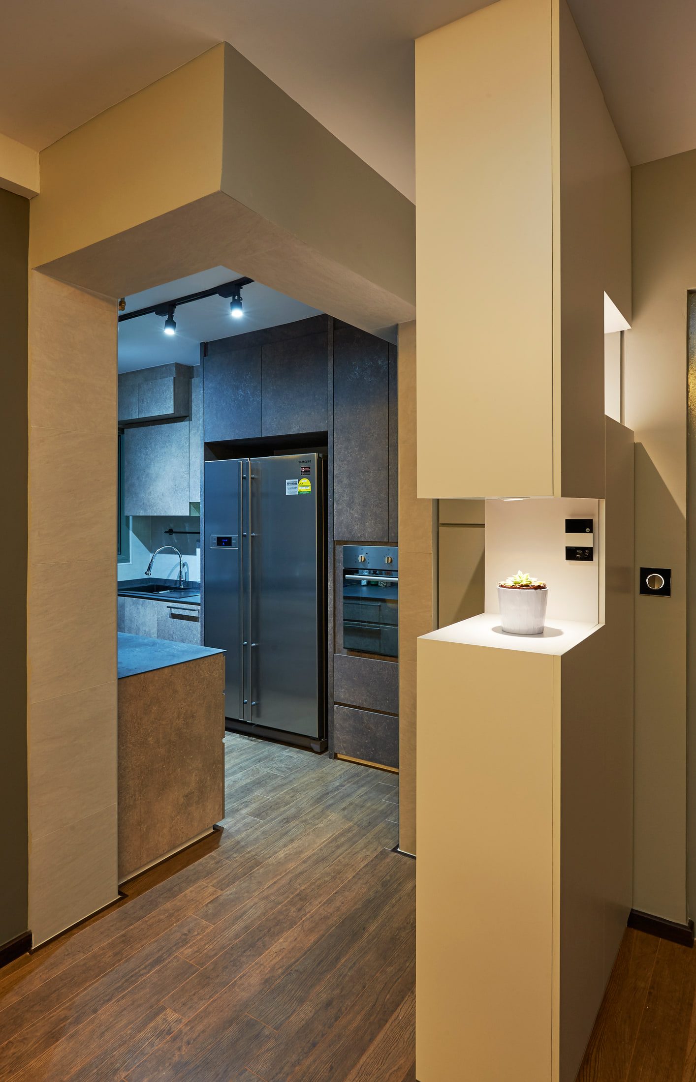 A doorway into the kitchen framed by pale joinery, with a stainless side-by-side fridge and built-in ovens beyond, and a lit display recess holding a single potted succulent.
