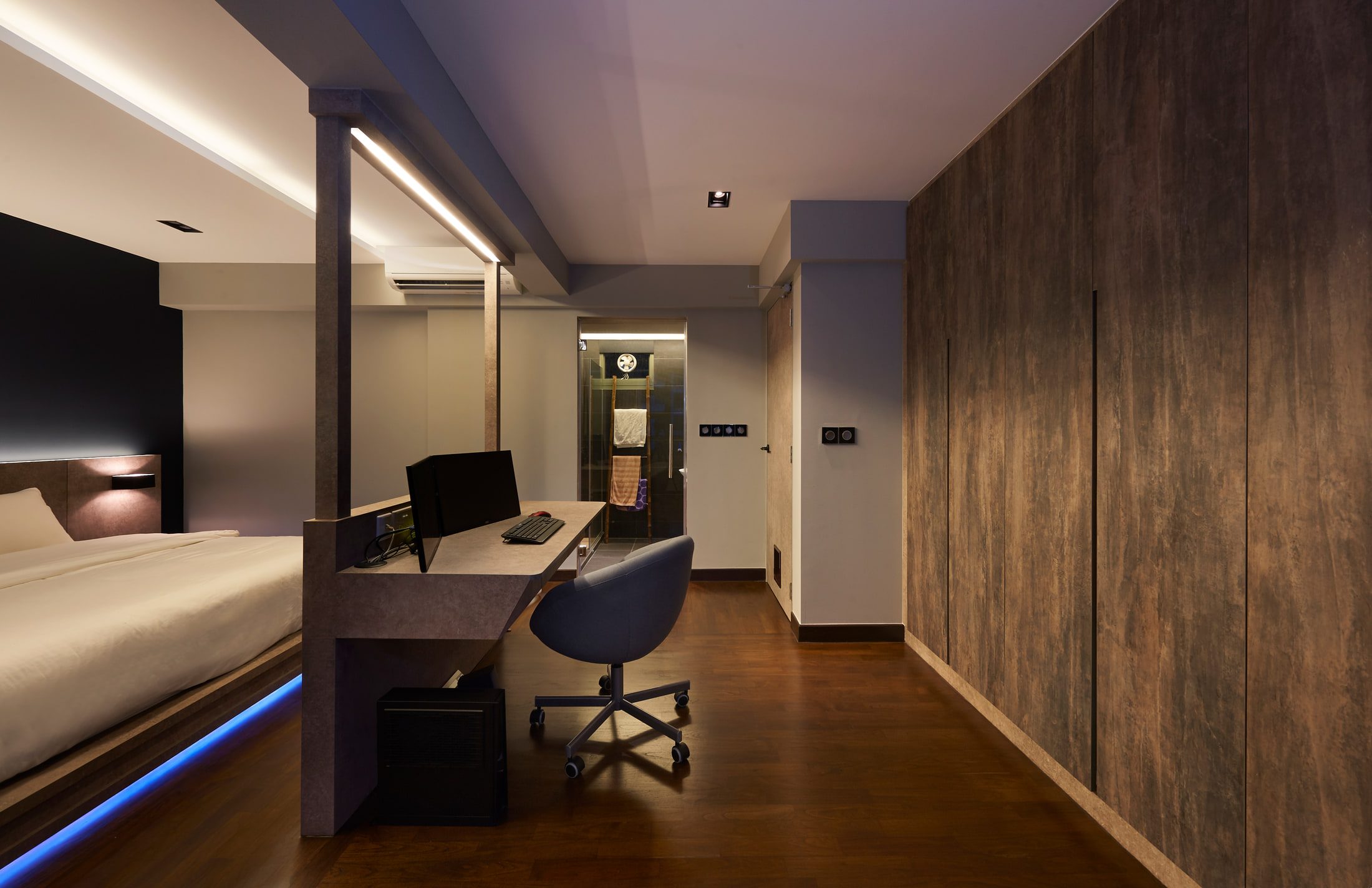 A bedroom seen from the wardrobe end at dusk, with a desk and monitor built into the stone-effect spine wall, a line of blue light under the bed platform, and the shower room lit beyond.