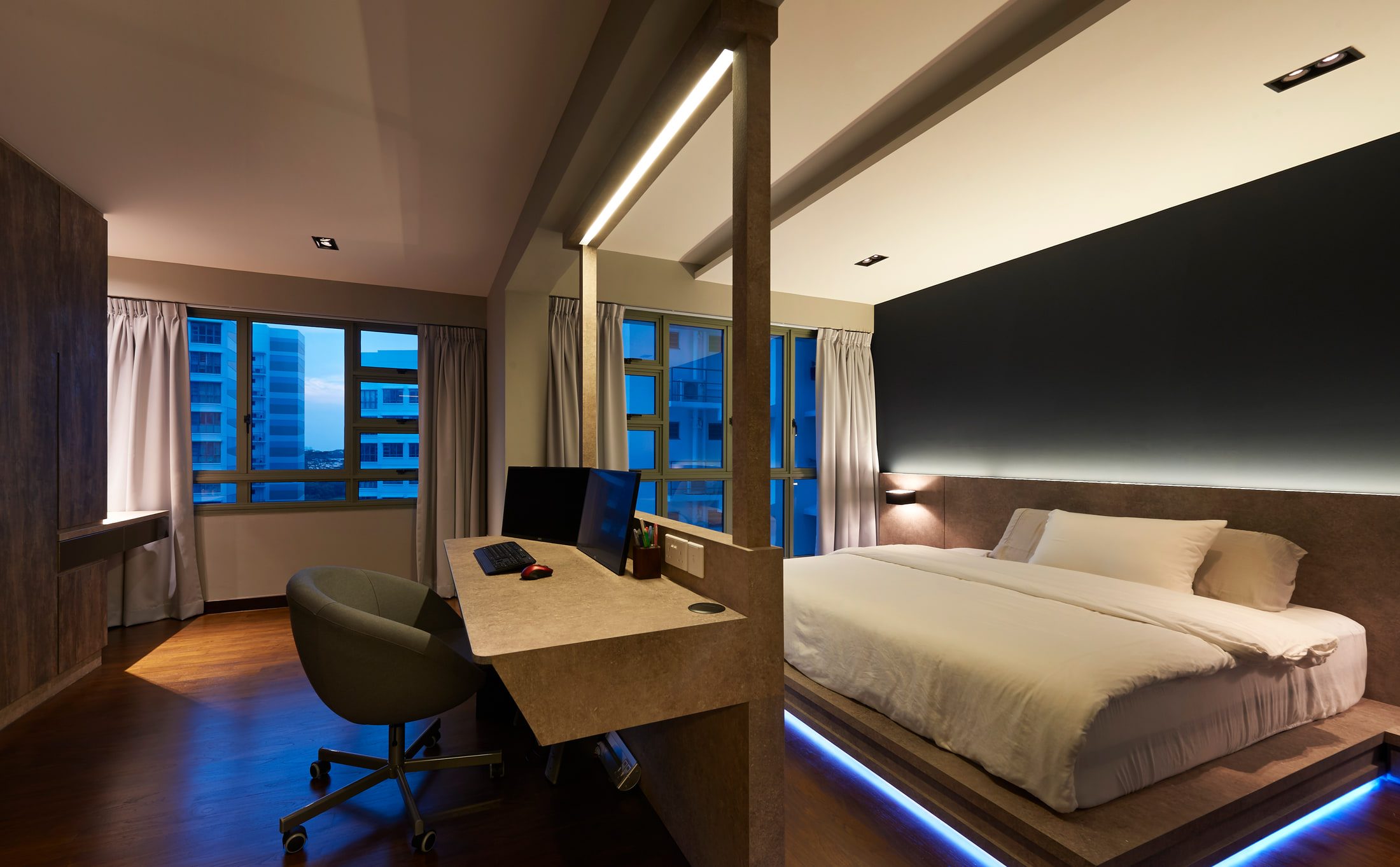 A bedroom at dusk with a desk built into a stone-effect spine wall, a low bed platform lit from underneath in blue, and a matt black wall behind the headboard.