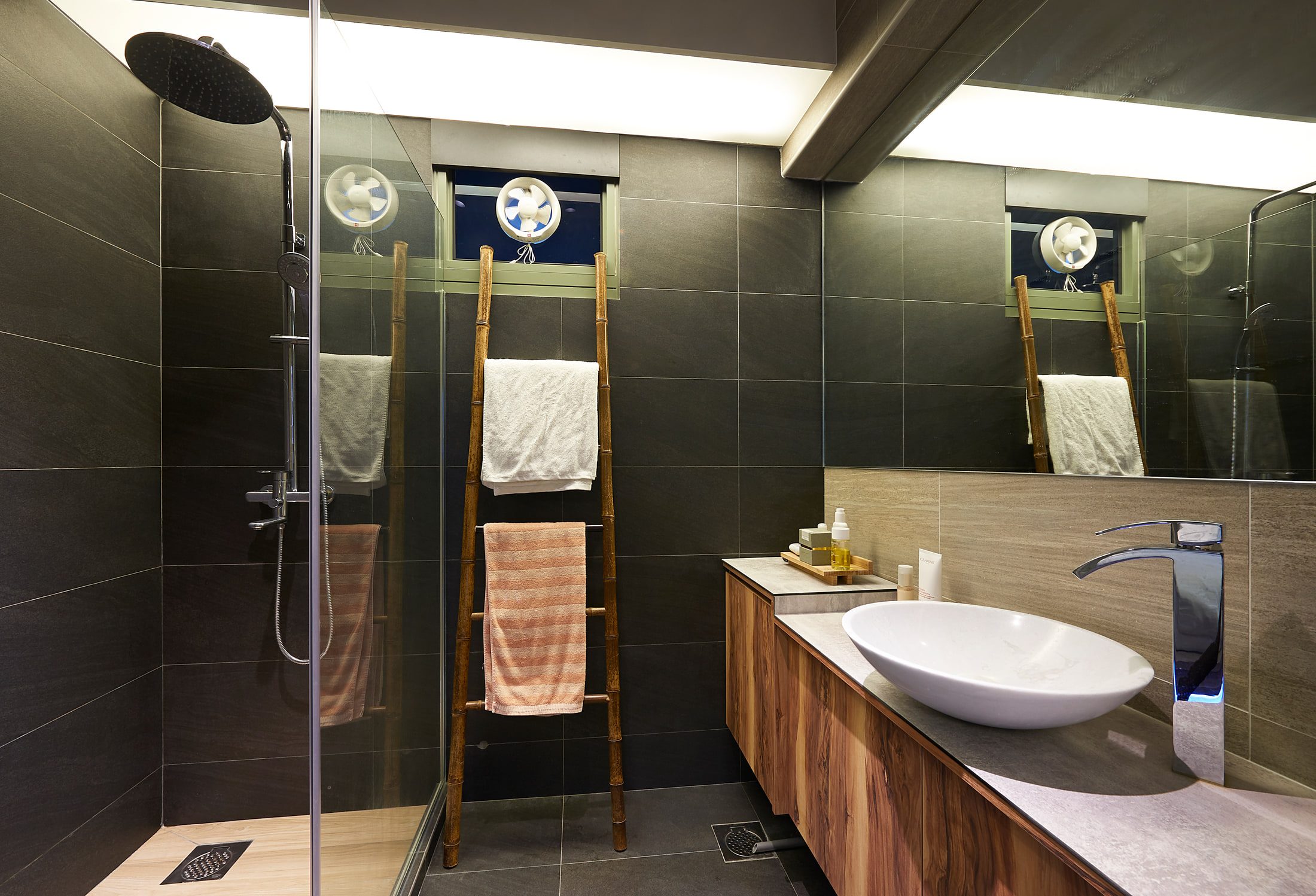A shower room in dark grey tile with a rain head behind a glass screen, a bamboo ladder hung with towels, and a white basin on a stone top over walnut drawers.