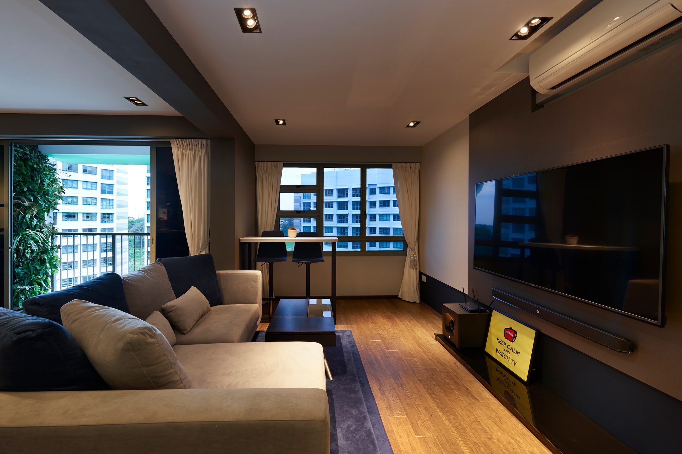 A living room with a grey and taupe sectional on a navy rug, a wall-hung television on a dark panelled wall, and a planted green wall on the balcony beyond.