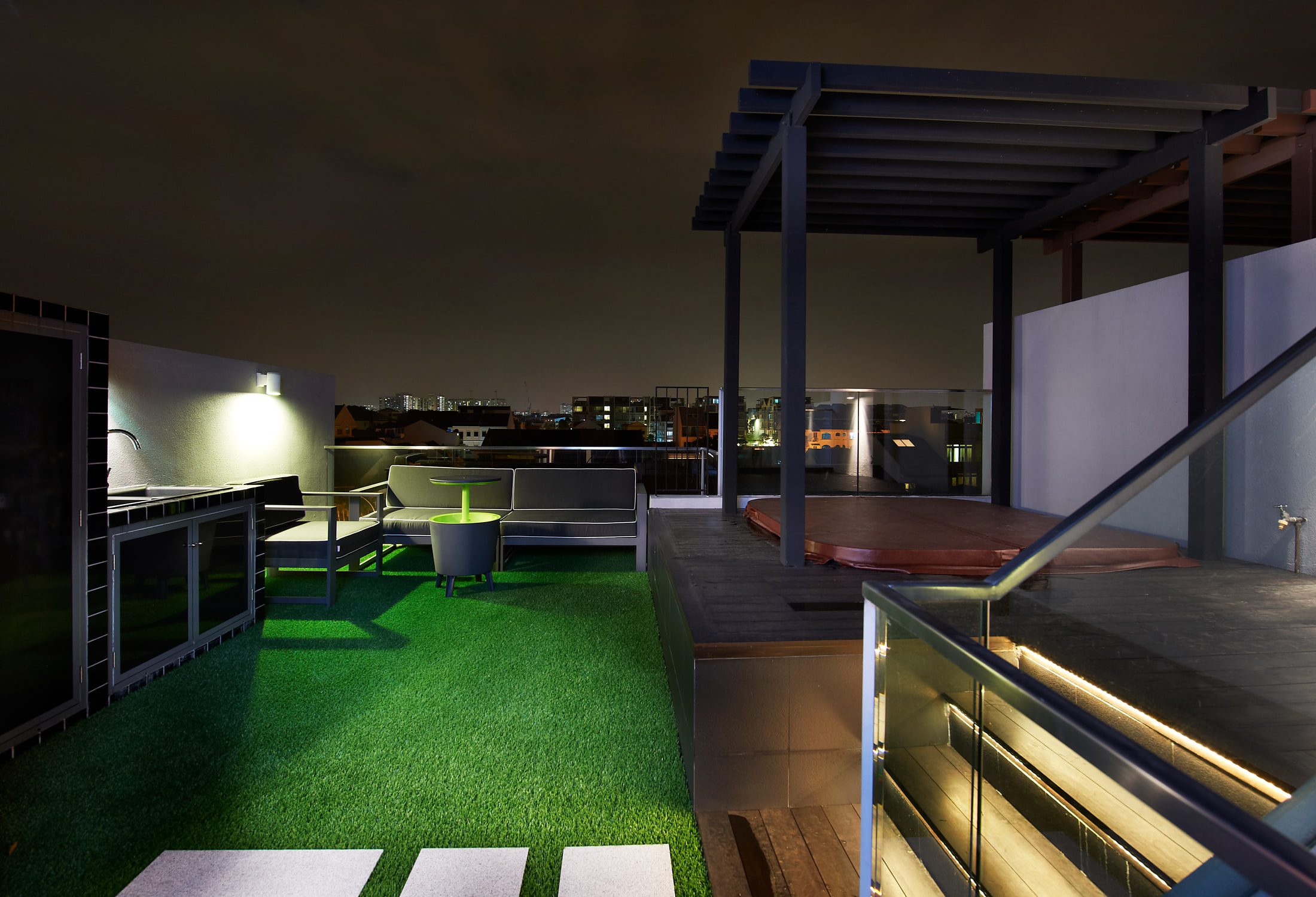 A roof terrace at night with artificial turf and a timber deck under a steel pergola, a covered spa, a low outdoor sofa and a green side table, and the city beyond a glass balustrade.