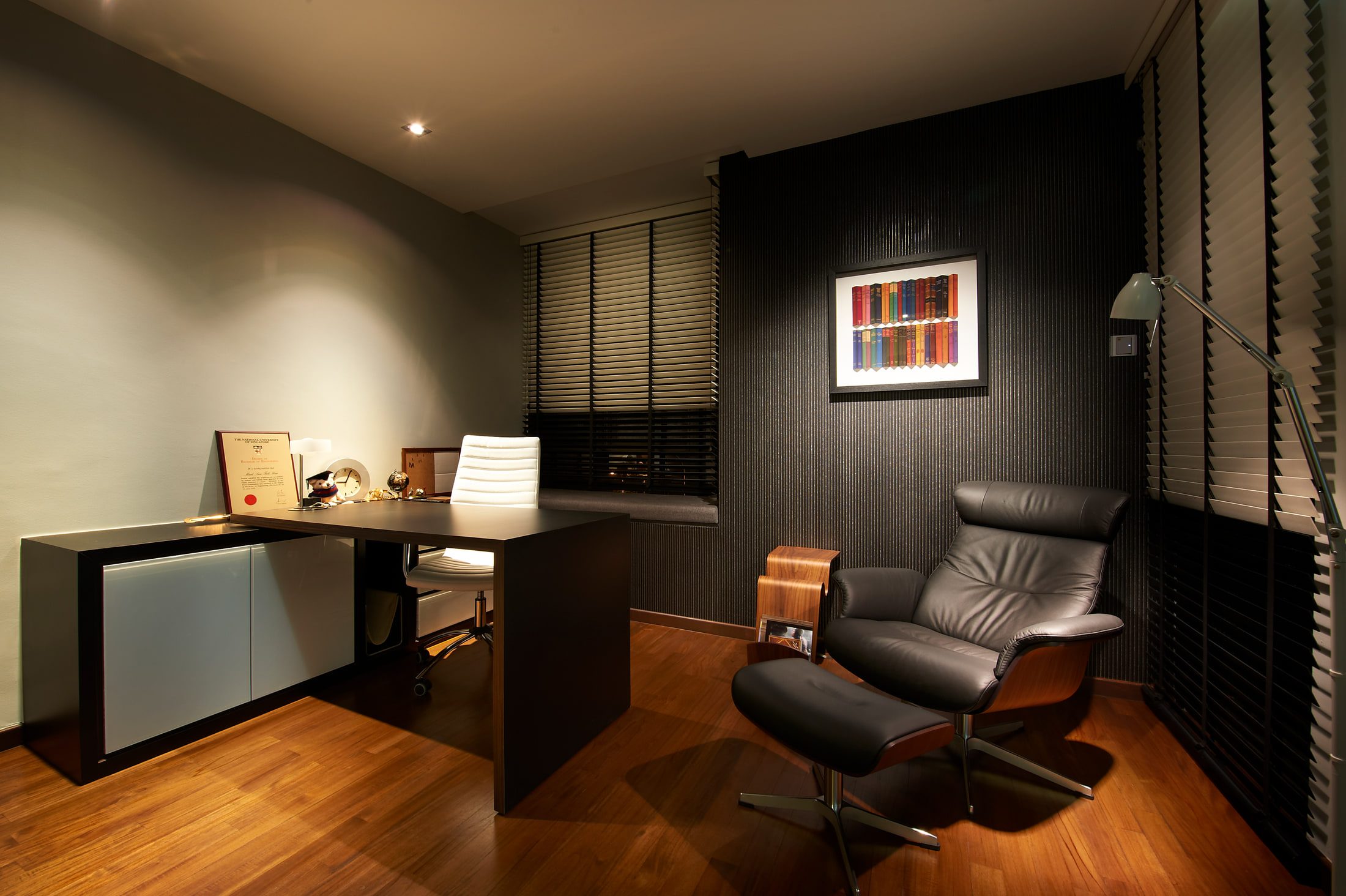 A study with a dark timber desk and white chair, a leather lounge chair and ottoman, and framed art on a black textured wall.