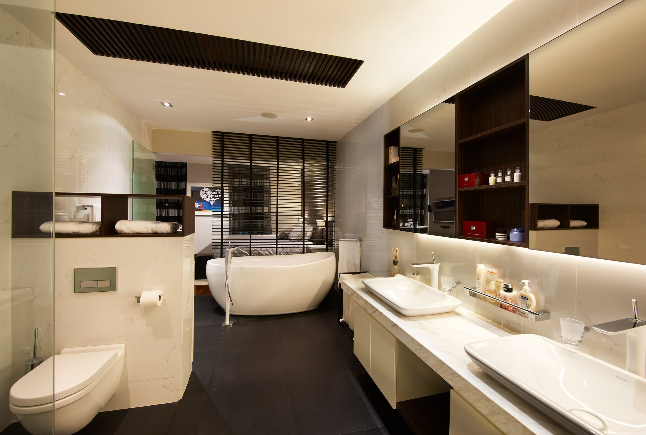An ensuite in white marble with a freestanding oval tub, twin basins on a marble ledge, a lit mirror band and dark stone floor, screened from the bedroom by black louvres.