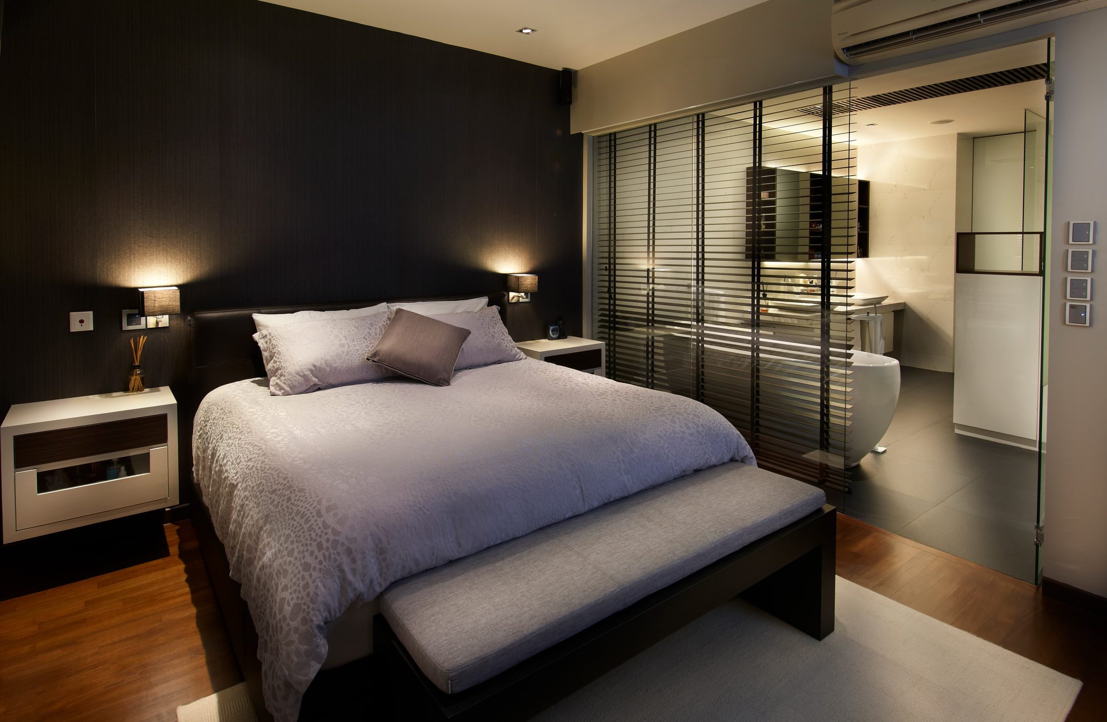 A bedroom with a dark textured headboard wall, wall-mounted reading lamps and a grey upholstered bench, opening through black timber louvres to a lit ensuite.