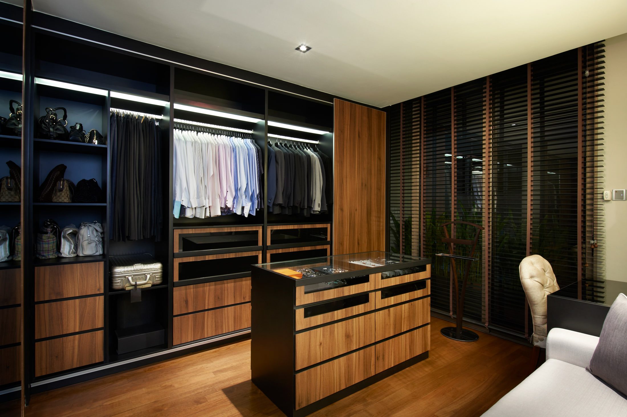 A walk-in wardrobe in black frame and walnut, with lit rails of shirts and suits, an island of drawers under a glass-topped watch display, and handbags on open shelves.