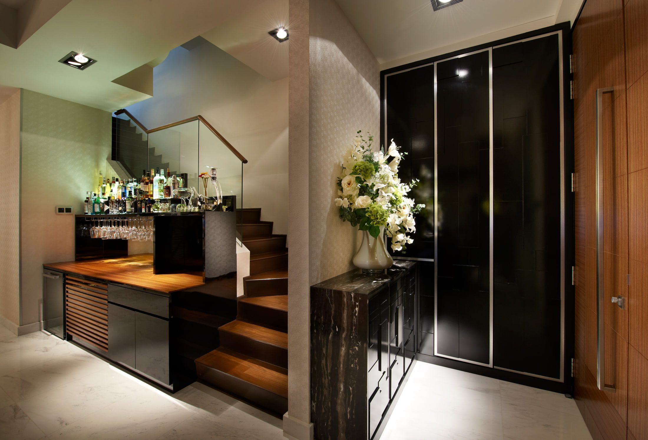 An entry hall with a home bar under a timber stair, glassware racked beneath the counter, and a black lacquer full-height cabinet beside a dark marble console holding white lilies.