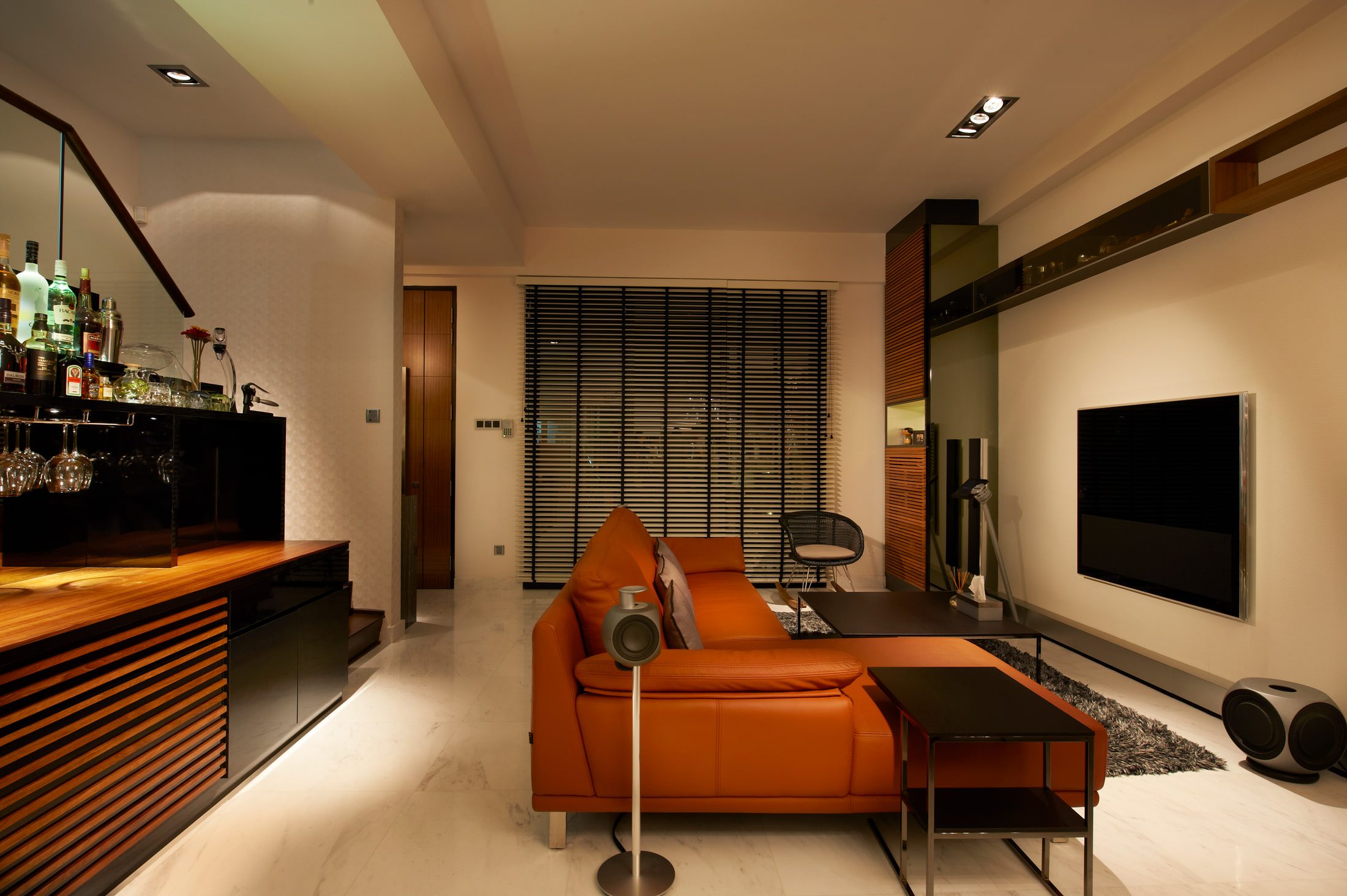 A living room with a tan leather sectional on white marble, a wall-mounted flat screen, and a home bar with a timber top and racked stemware in the foreground.