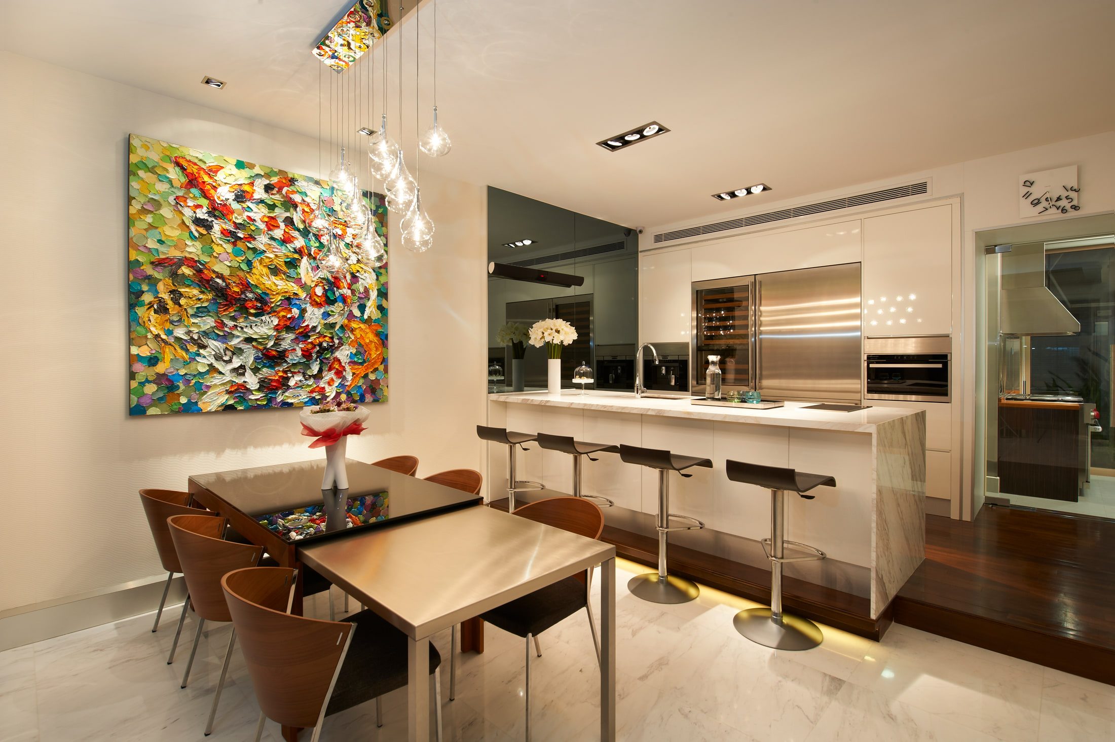 A dining and kitchen room in white marble, with a palette-knife koi painting, a cluster of clear glass drop pendants over a dark glass table, and a marble-clad island backed by a stainless appliance wall.