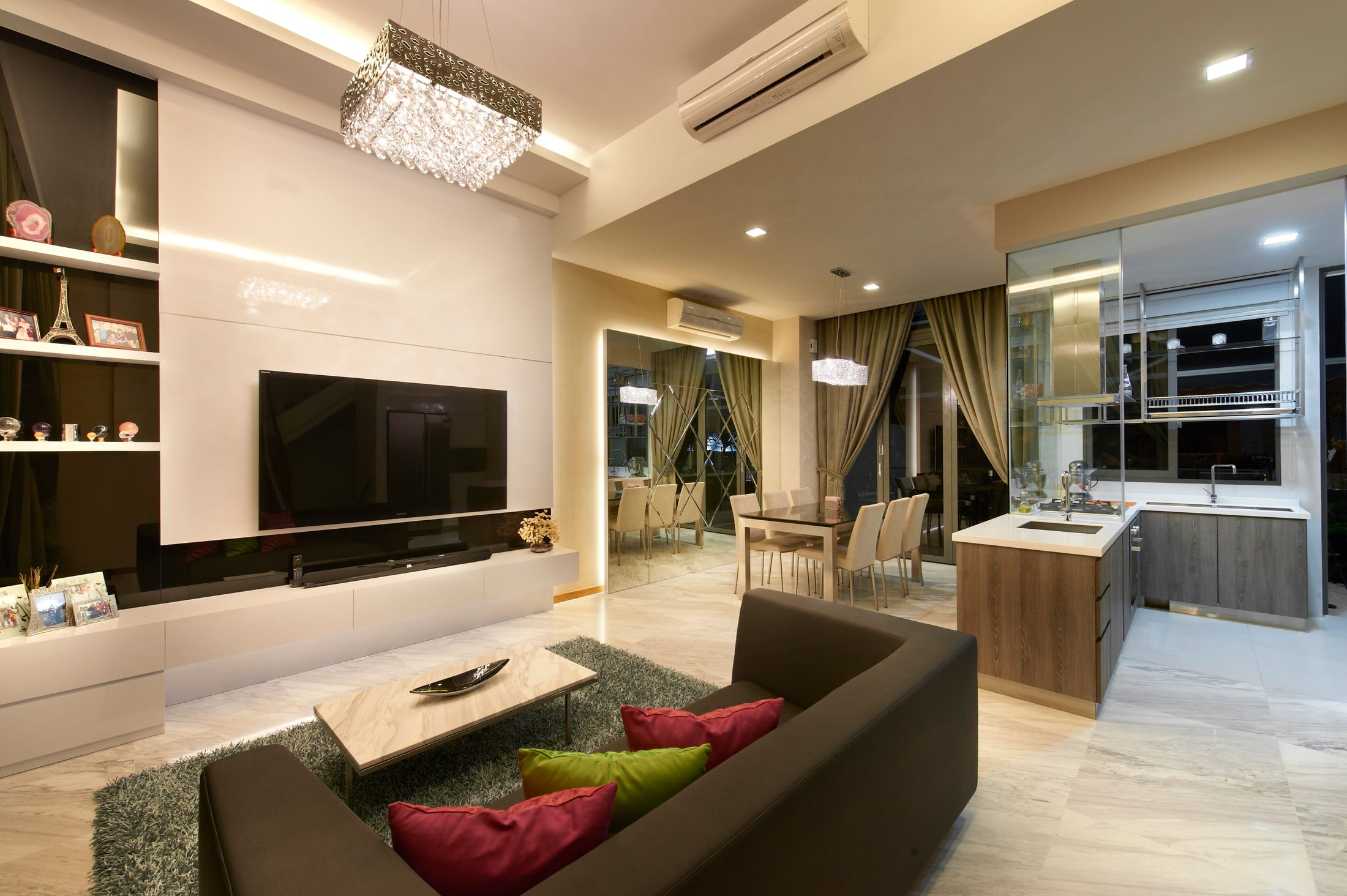 An open living, dining and kitchen floor in pale marble, with a rectangular crystal chandelier over the sofa, a full-height mirrored return, and a glazed kitchen screen at the far end.