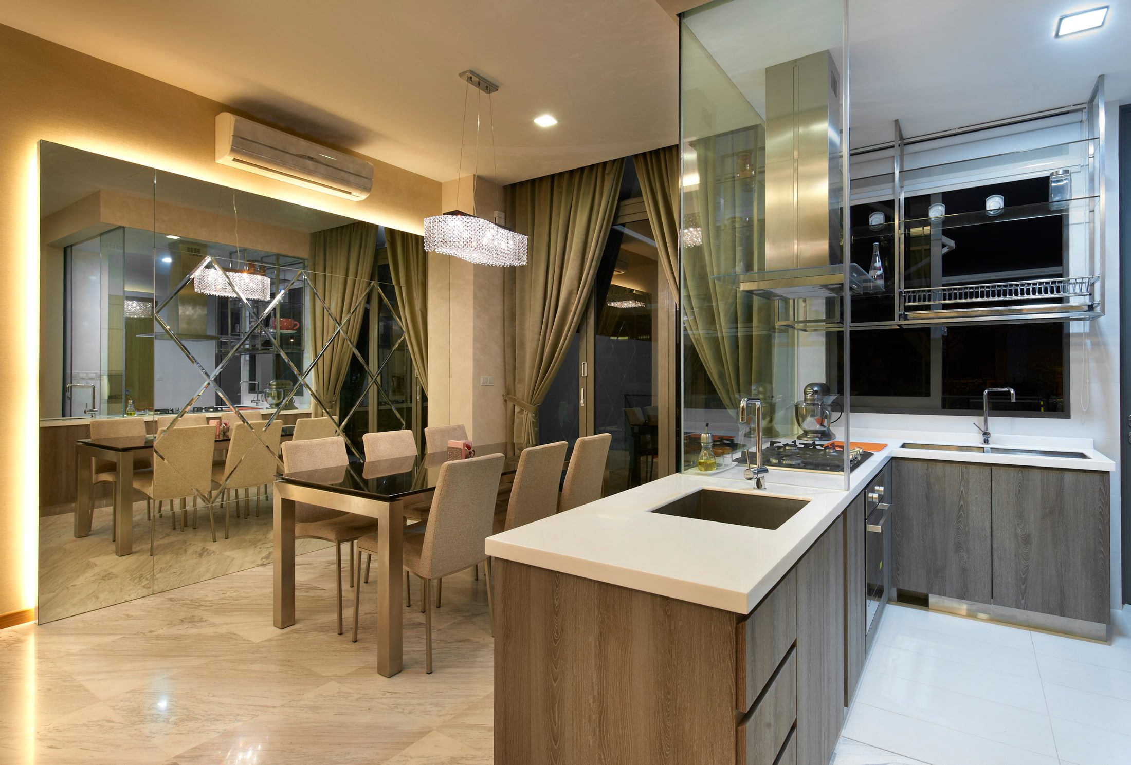 A dining table in bronze-framed glass with linen chairs beside a mirror wall set with bevelled diamond panels, and a kitchen island in grey oak under a white worktop.