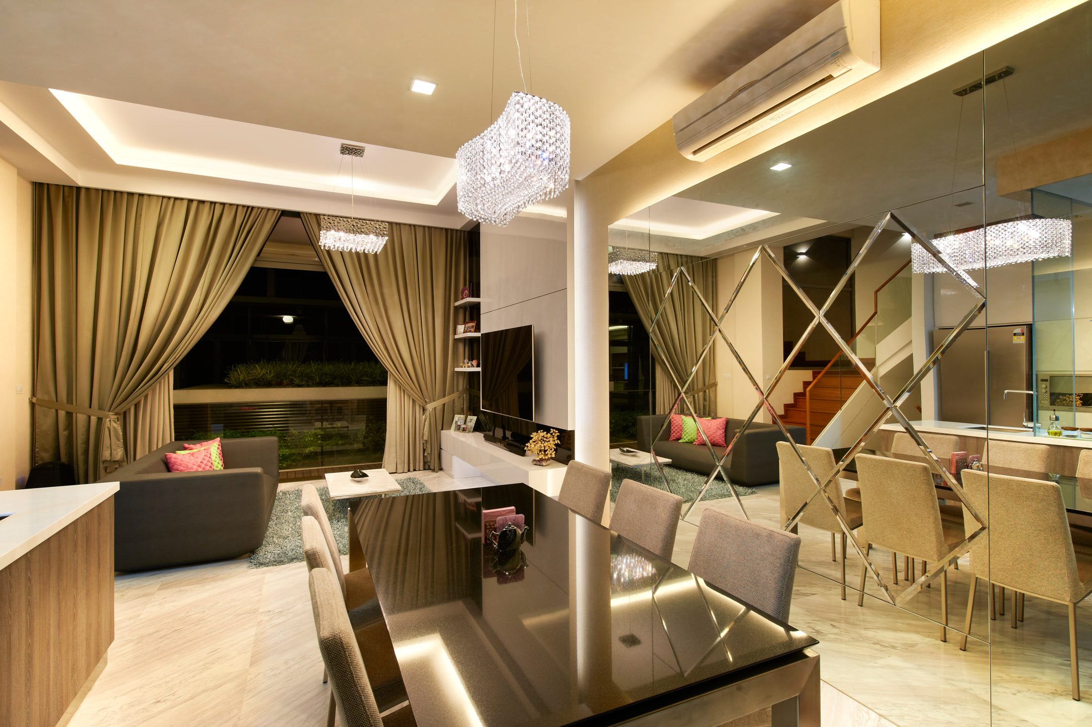 A living and dining room with a crystal chandelier over a glass table, gold curtains to full-height glazing and mirrored panels in a diamond lattice.