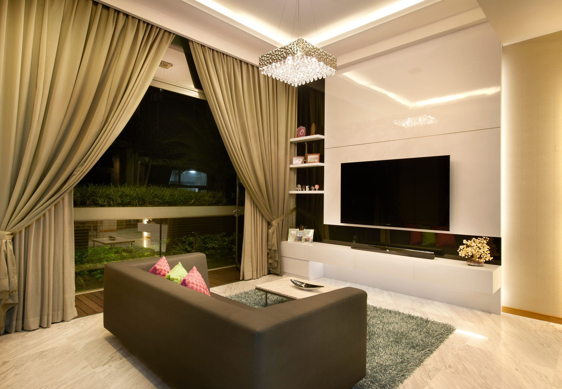 A living room in white marble with floor-length taupe drapery drawn back from a garden window, a gloss white media wall with a slim black recess, and a crystal-drop chandelier over a charcoal sofa.