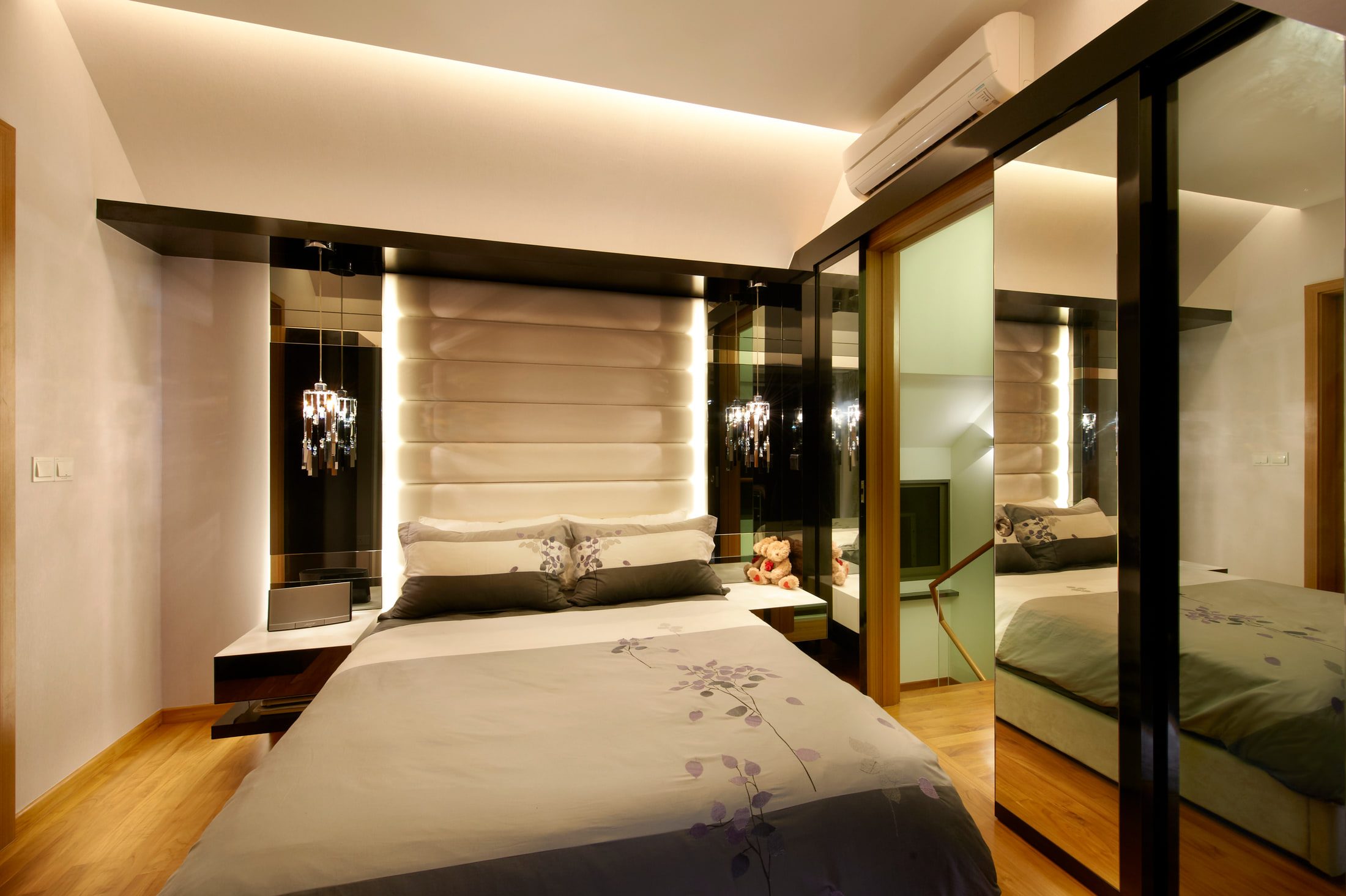 A bedroom with a tall padded headboard in cream leather lit from its edges, mirrored and glass panels either side, and a crystal drop pendant hanging over the bedside shelf.