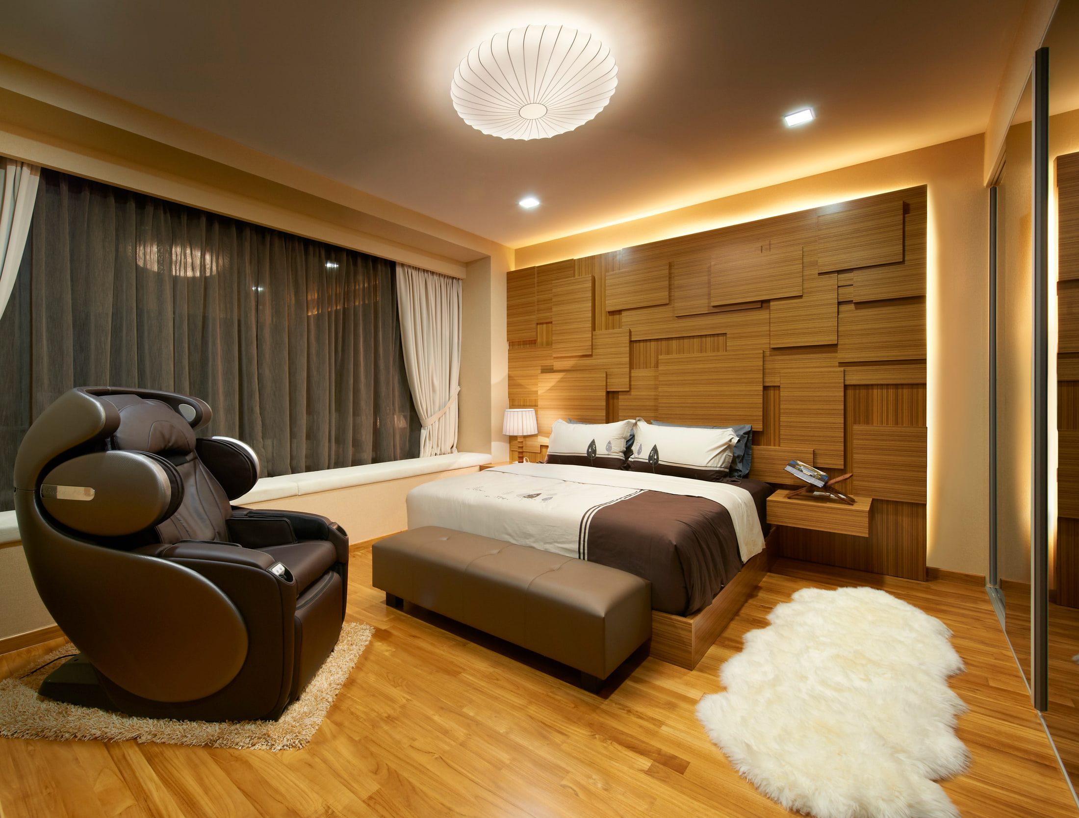A bedroom with a headboard wall of staggered timber blocks lit from behind, a brown leather recliner on a shaggy rug, and a sheepskin beside the mirrored wardrobe run.