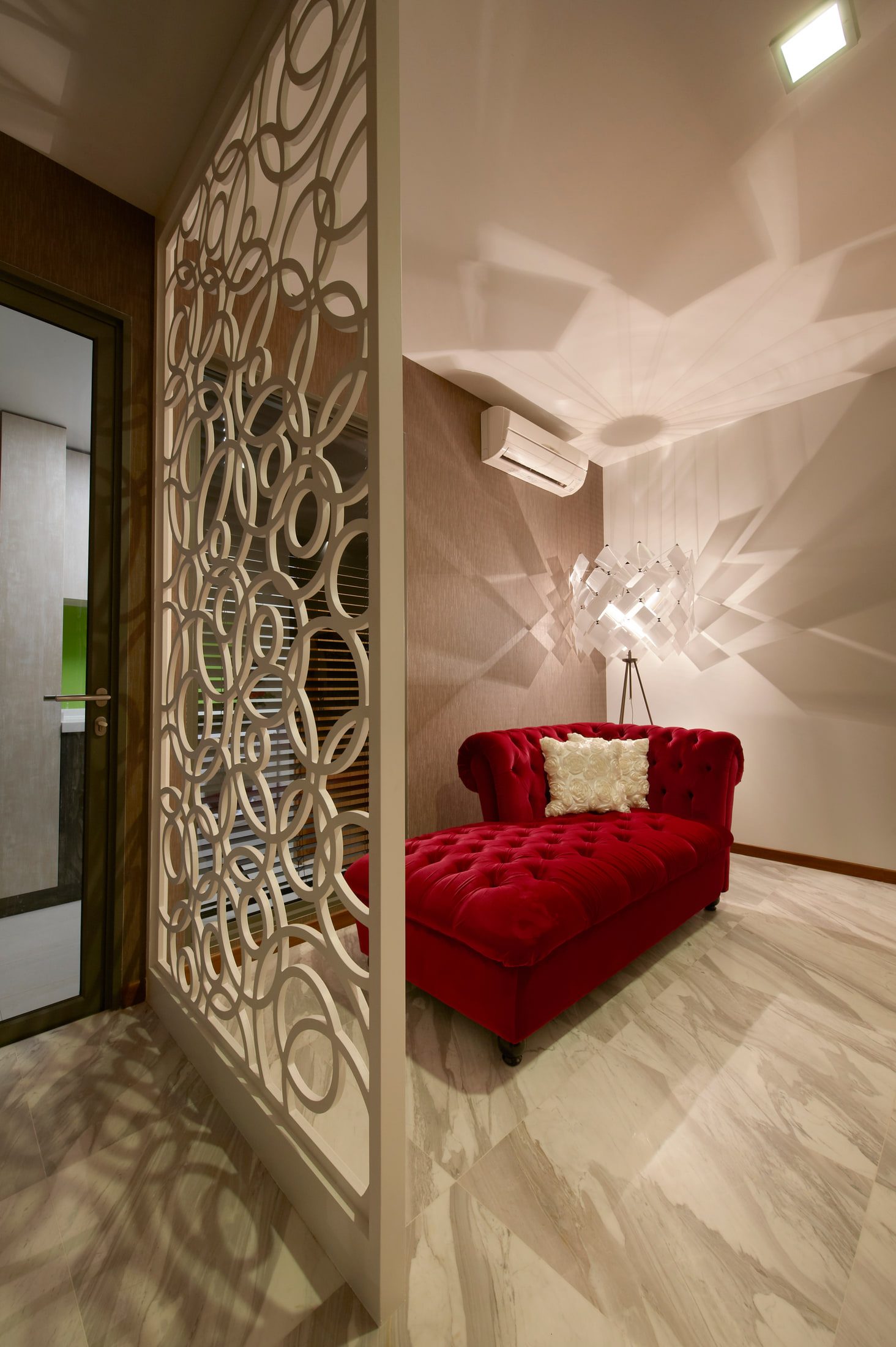 A red buttoned velvet chaise on veined marble beneath a faceted acrylic pendant, screened from the entrance by a cream fretwork panel of interlocking circles.