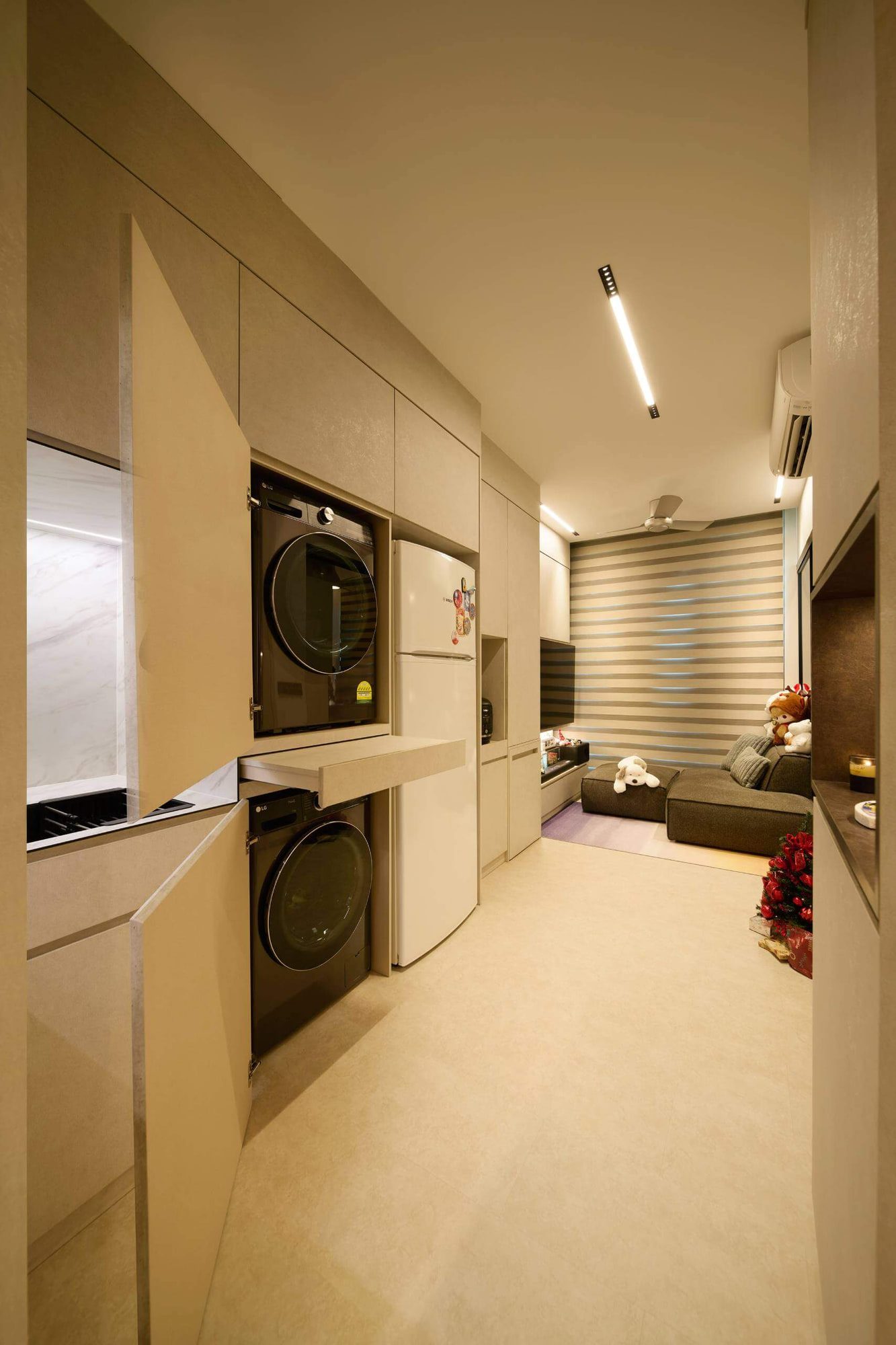 A galley kitchen with a marble-veined splashback faces a run of flush pale joinery concealing laundry appliances, opening onto a living area with low modular seating.