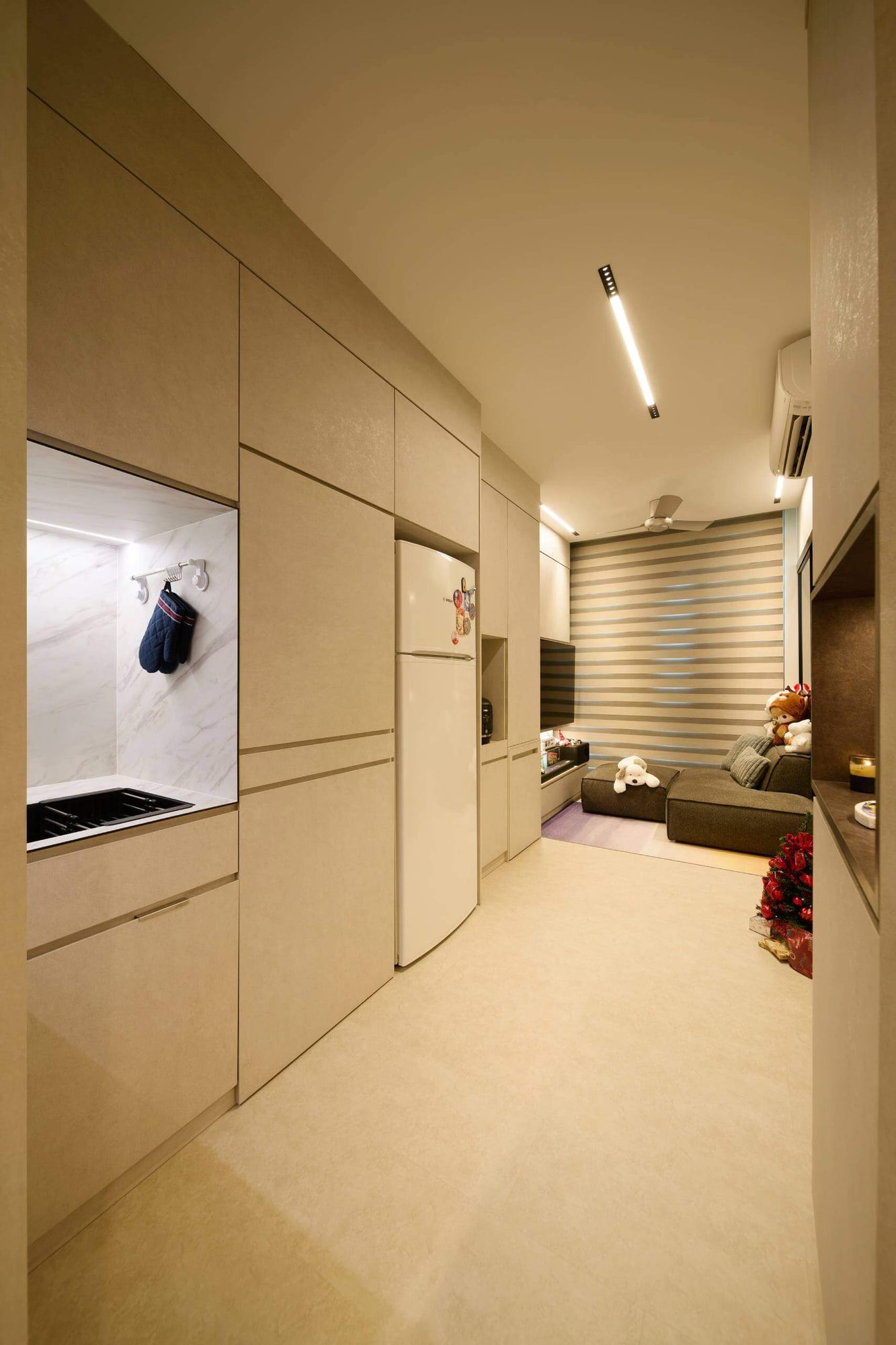 A corridor lined in cream joinery with lit display niches, a concealed kitchenette at left and the living area beyond.