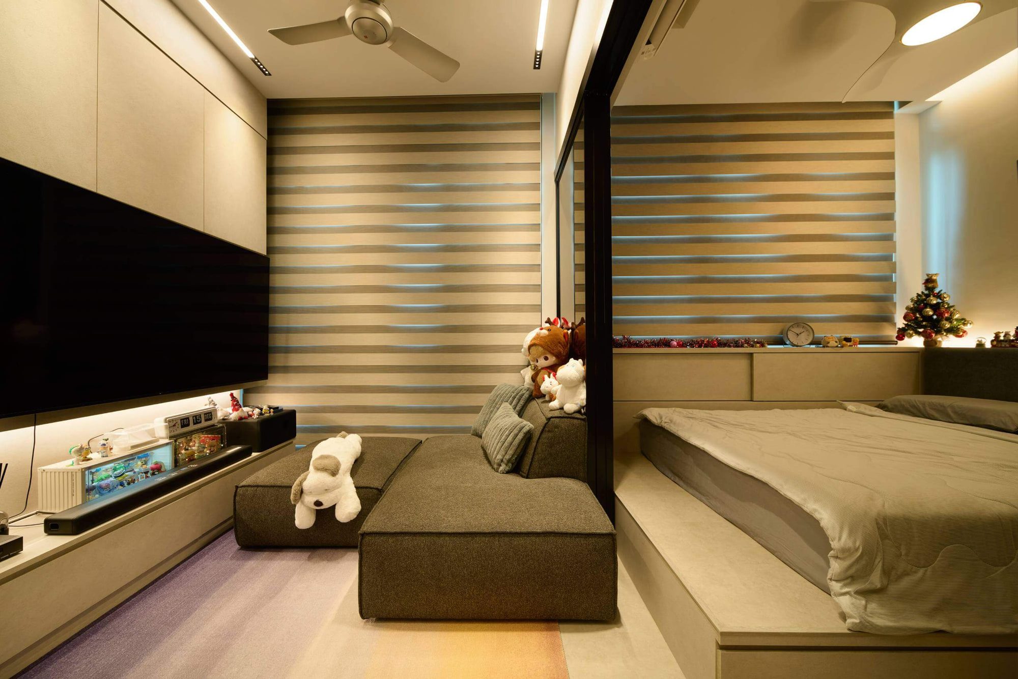 Low modular seating faces a wall-mounted screen on pale joinery, with a raised platform bed behind a bronze-tinted glass partition and blinds across two walls.