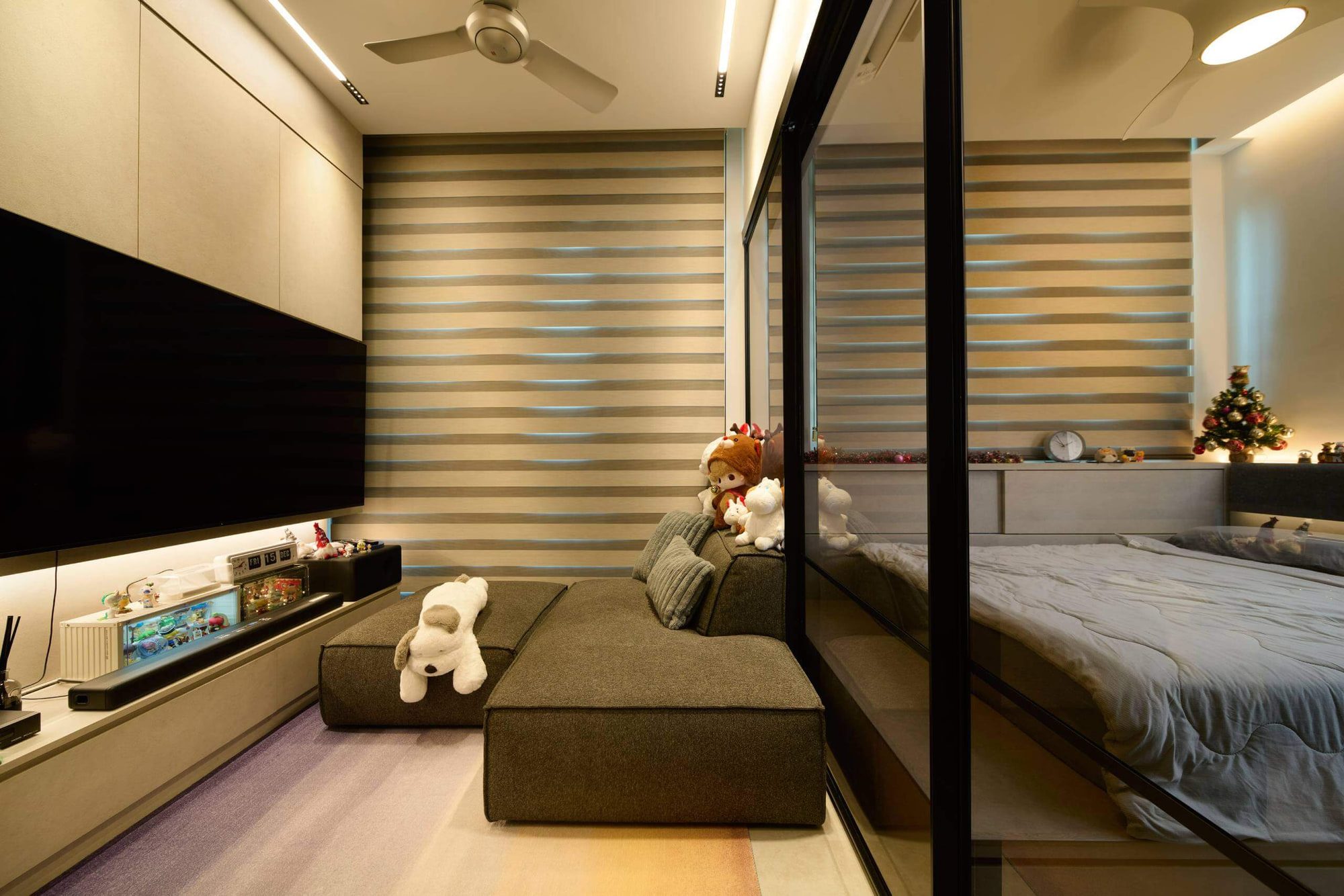 A bedroom with a slim television ledge, a striped roller blind at the window and a mirrored wardrobe returning along the bed.