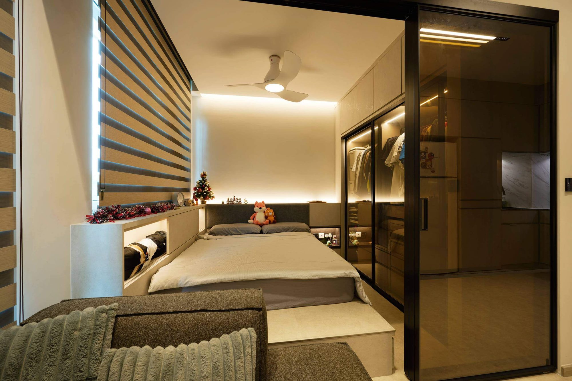A mattress on a pale joinery platform with an open storage ledge beneath the window, an upholstered headboard, overhead cabinets and tinted glass wardrobe doors.