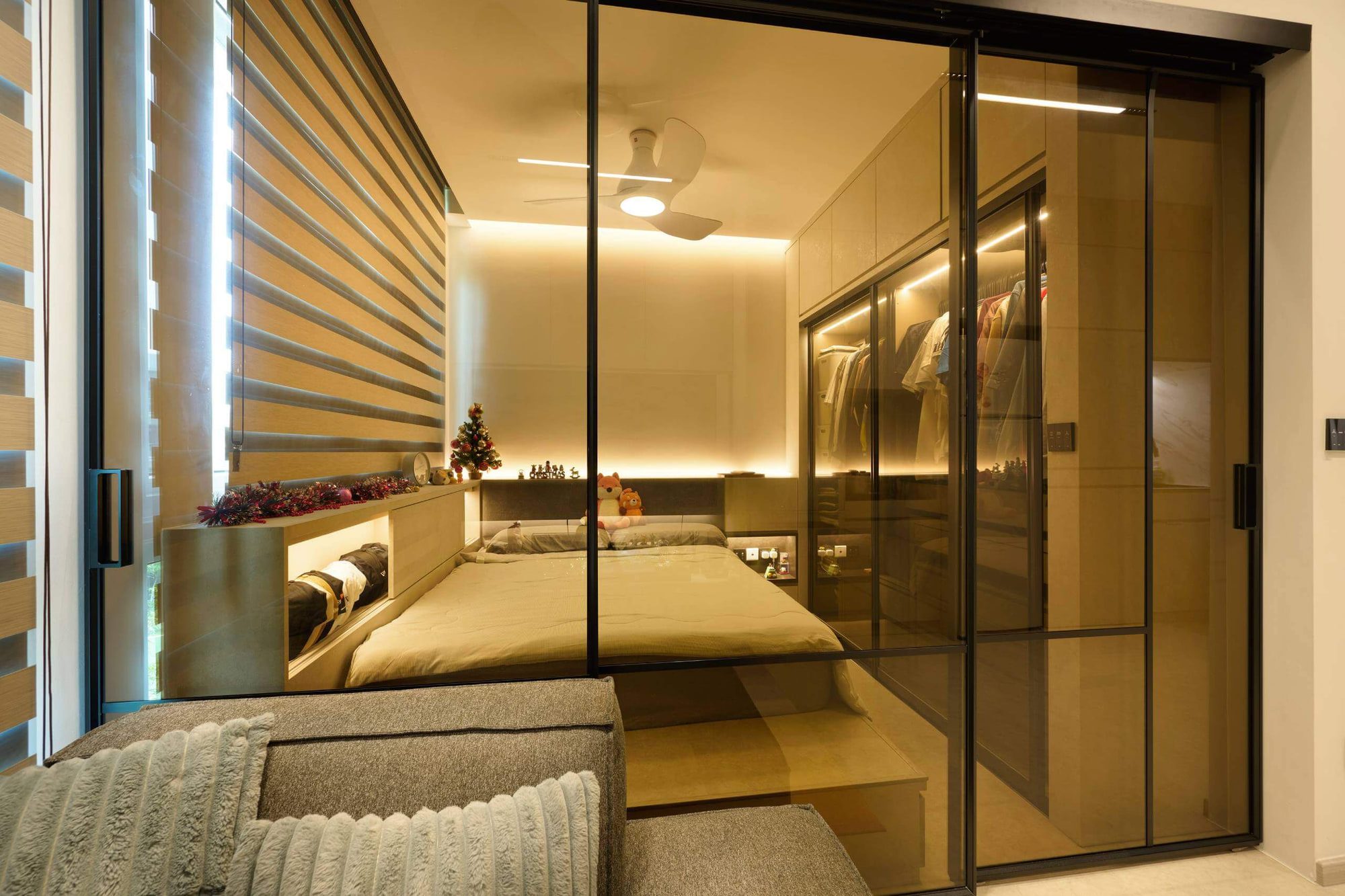 A bedroom with a platform bed against a striped blind, enclosed by bronze-tinted glass sliding doors to the wardrobe.
