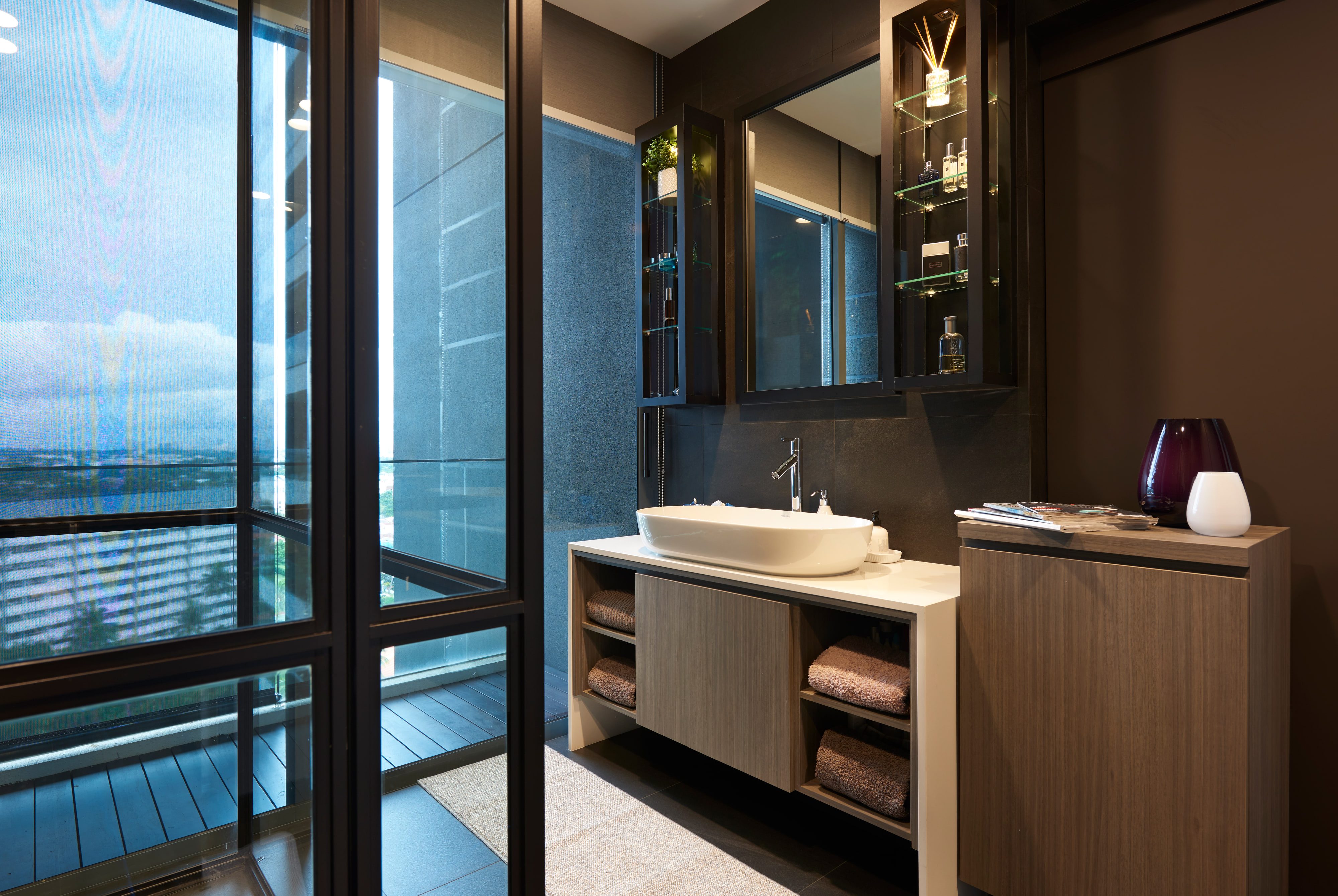 A dark-toned bathroom with a white vessel basin on a pale timber vanity, mirrored cabinets above, and a glazed door opening to a narrow balcony.