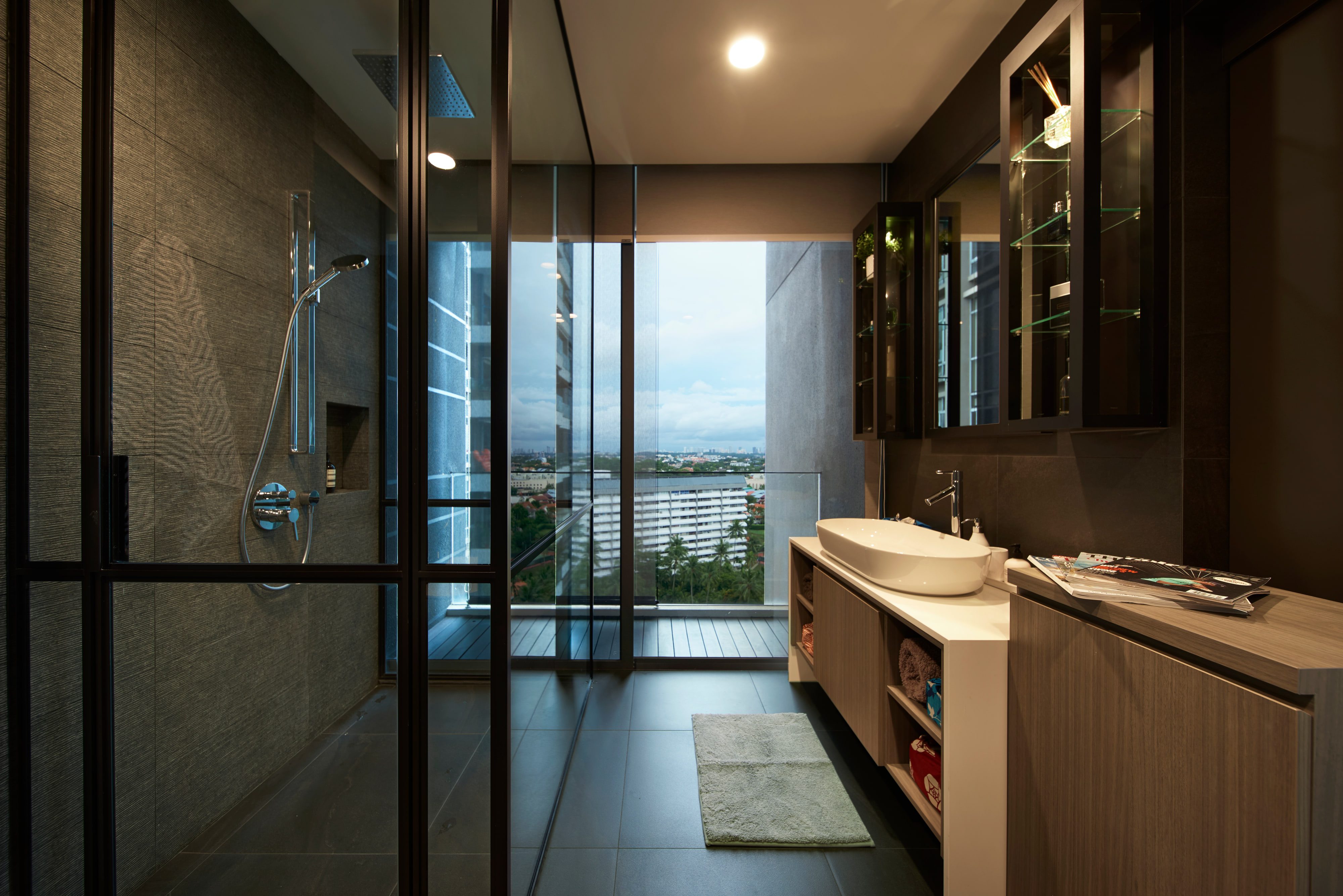 A walk-in shower behind a black-framed glass screen faces a wall-hung vanity across charcoal stone tile, with daylight entering from the balcony glazing beyond.