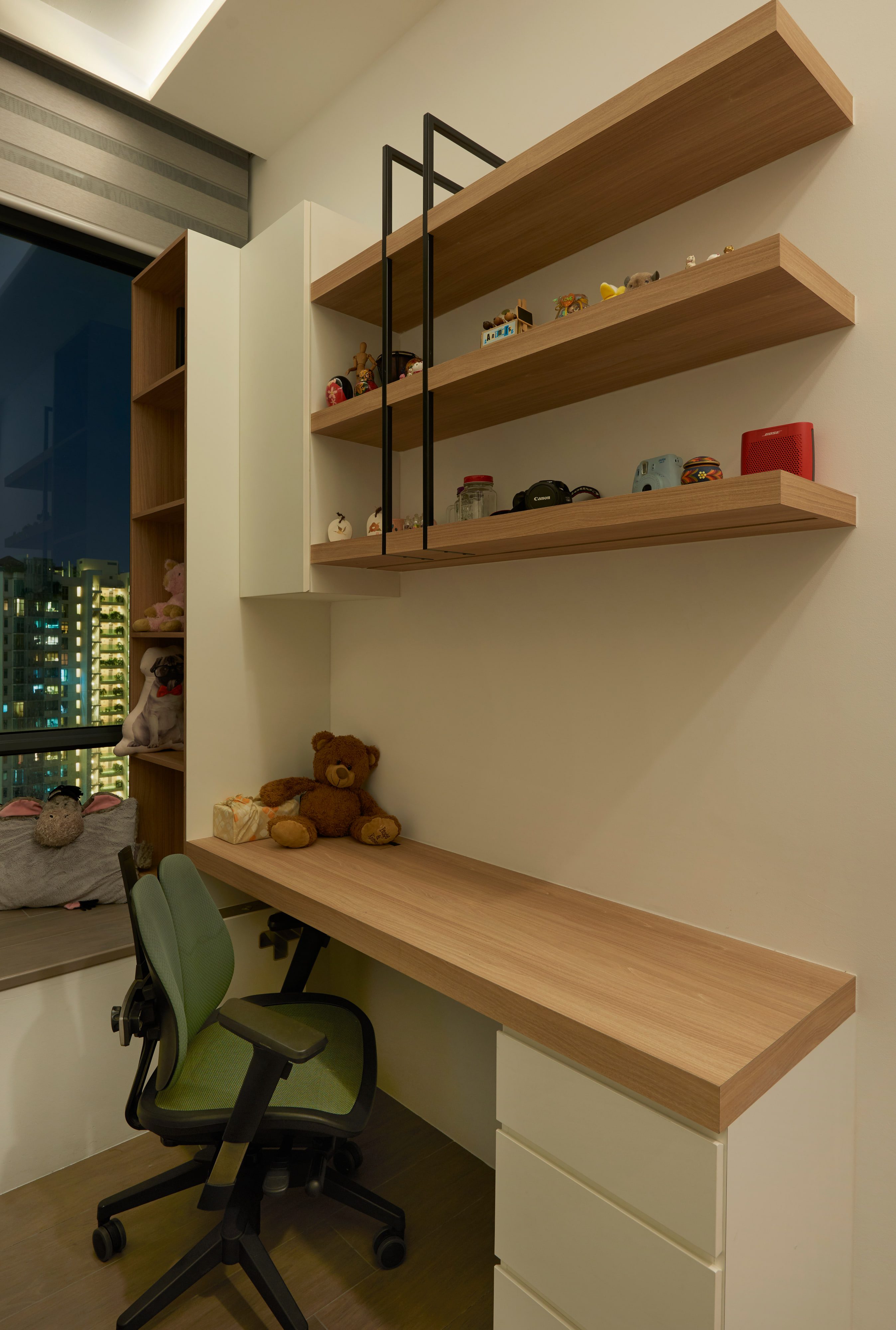 A cantilevered timber desk with staggered floating shelves crossed by slim black uprights, a white drawer unit below and a cushioned window bench alongside.