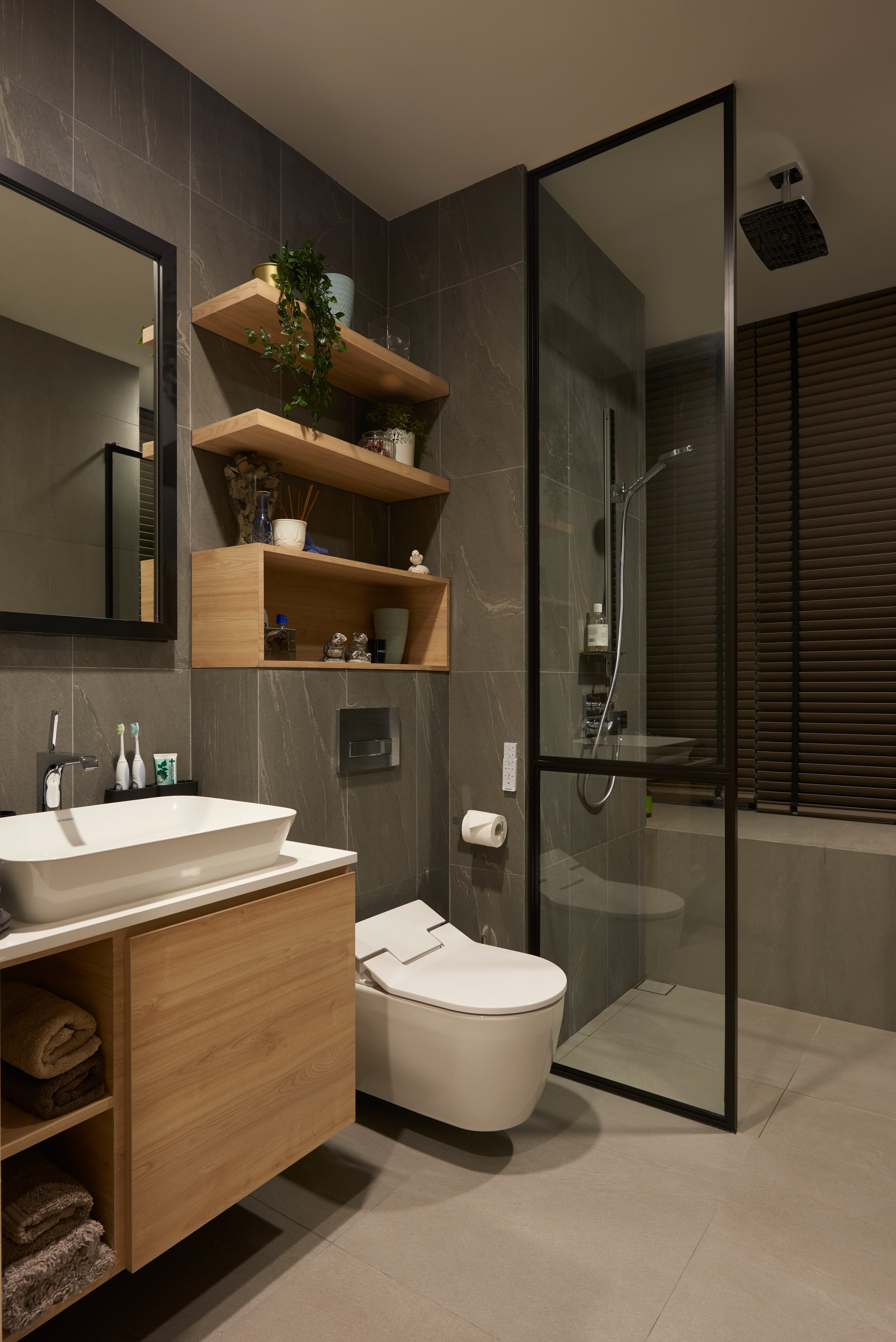 A bathroom with a timber wall-hung vanity and open shelving, a white basin on a stone top and a glazed shower against grey tile.