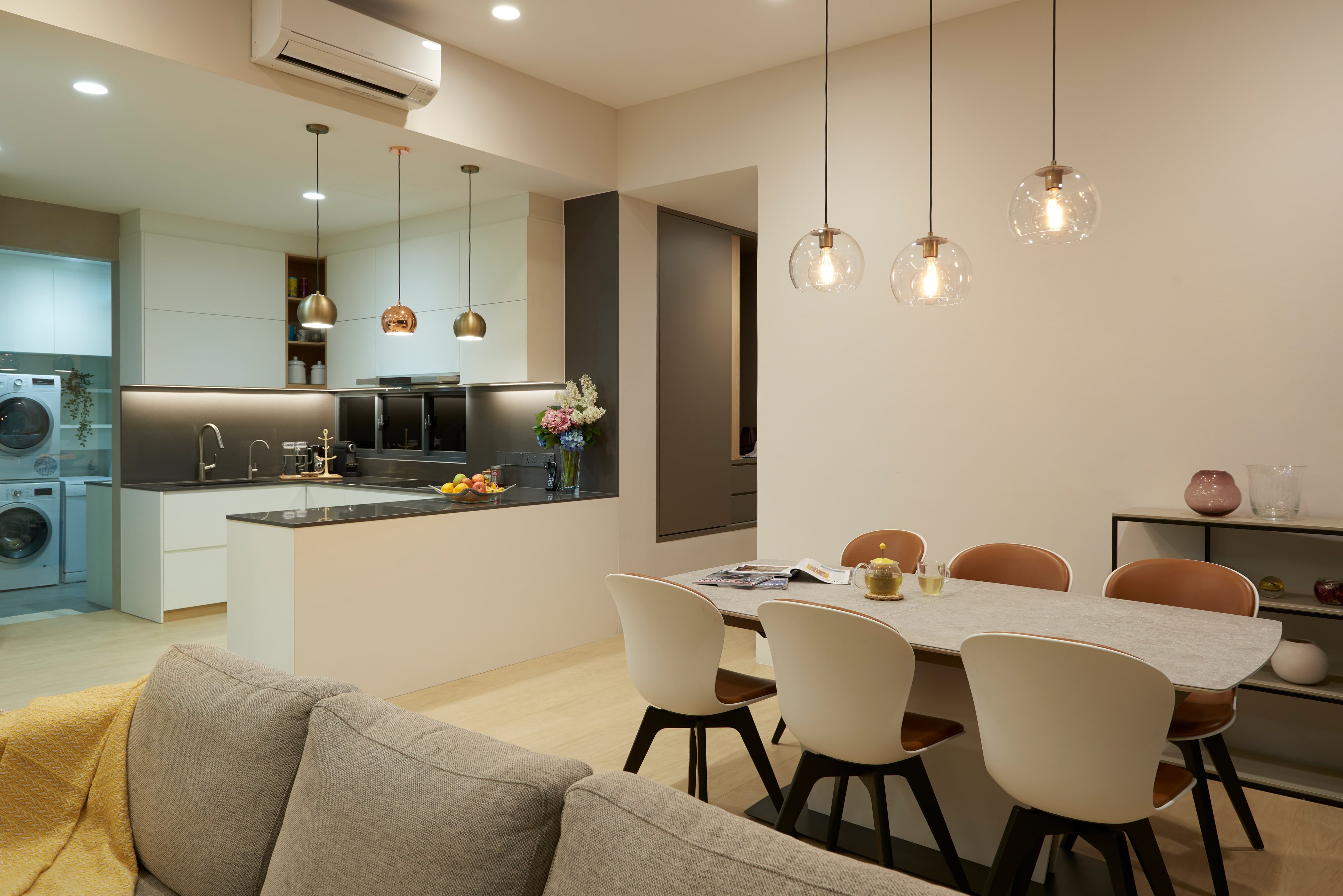 An open kitchen and dining room with three glass globe pendants over a pale table, a stone island and a utility bay beyond.