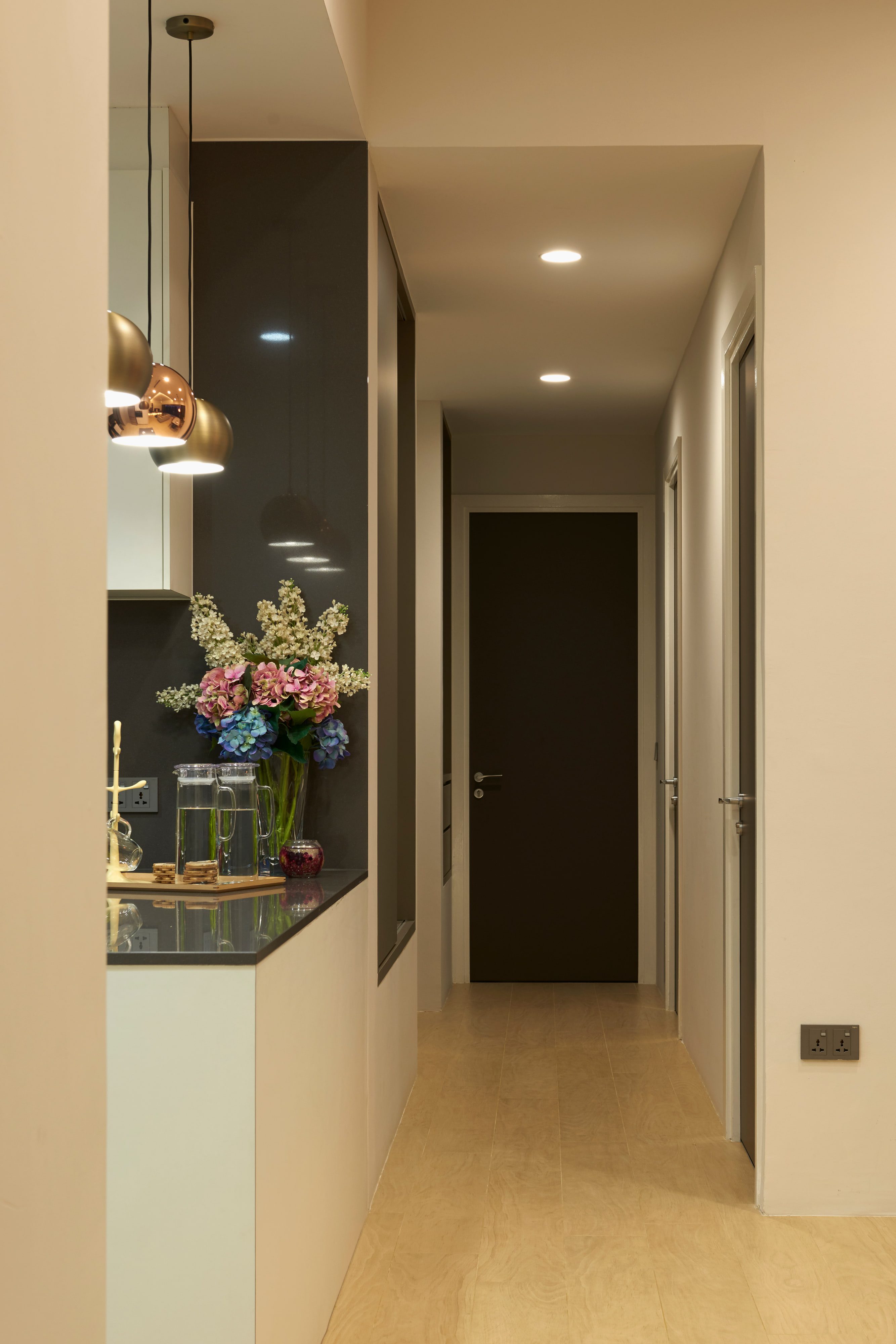 A hall running to a dark timber door, with a copper pendant over a stone counter and a vase of flowers standing on it.
