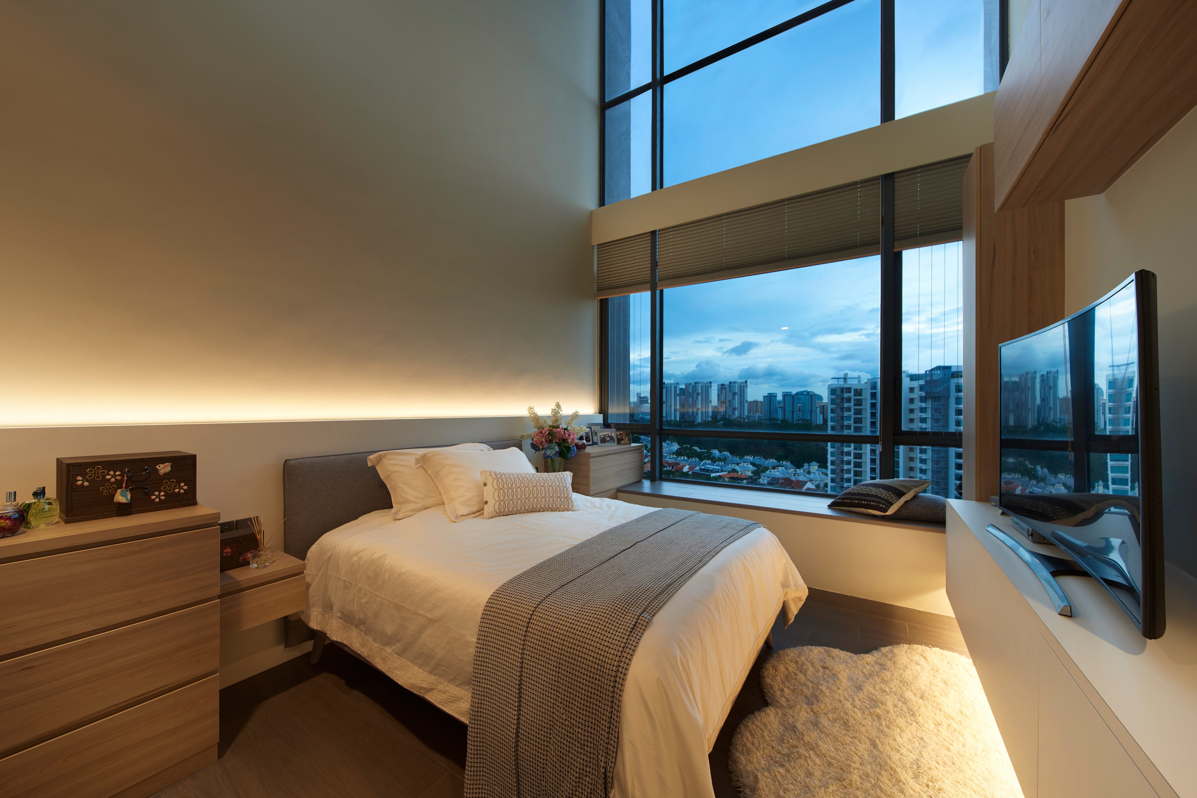 A bedroom with full-height corner glazing over the city, a timber-lined headboard wall and a cove-lit ceiling.