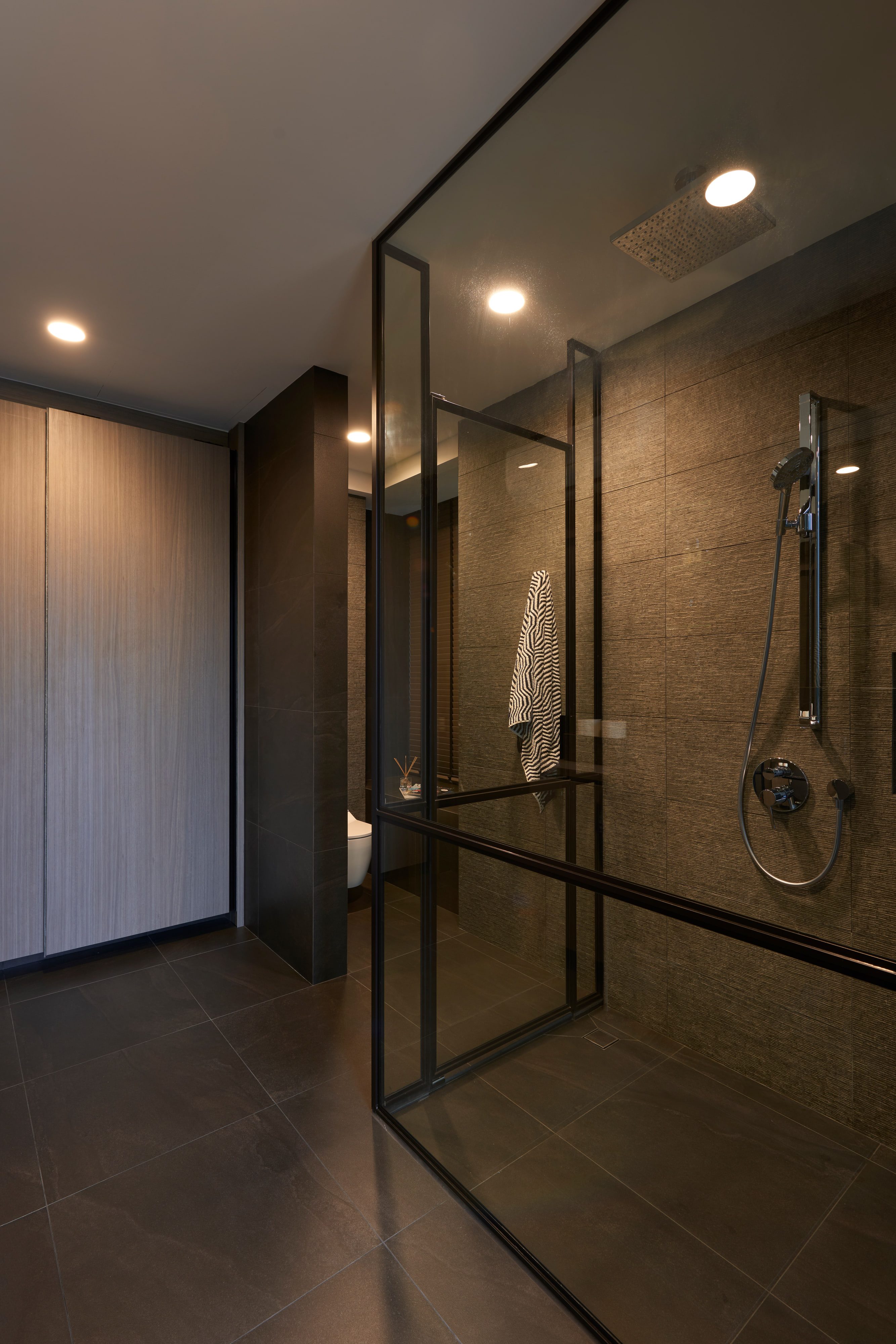 A shower room in dark stone behind a black-framed glass screen, with a rain head above and a timber-lined wall at left.
